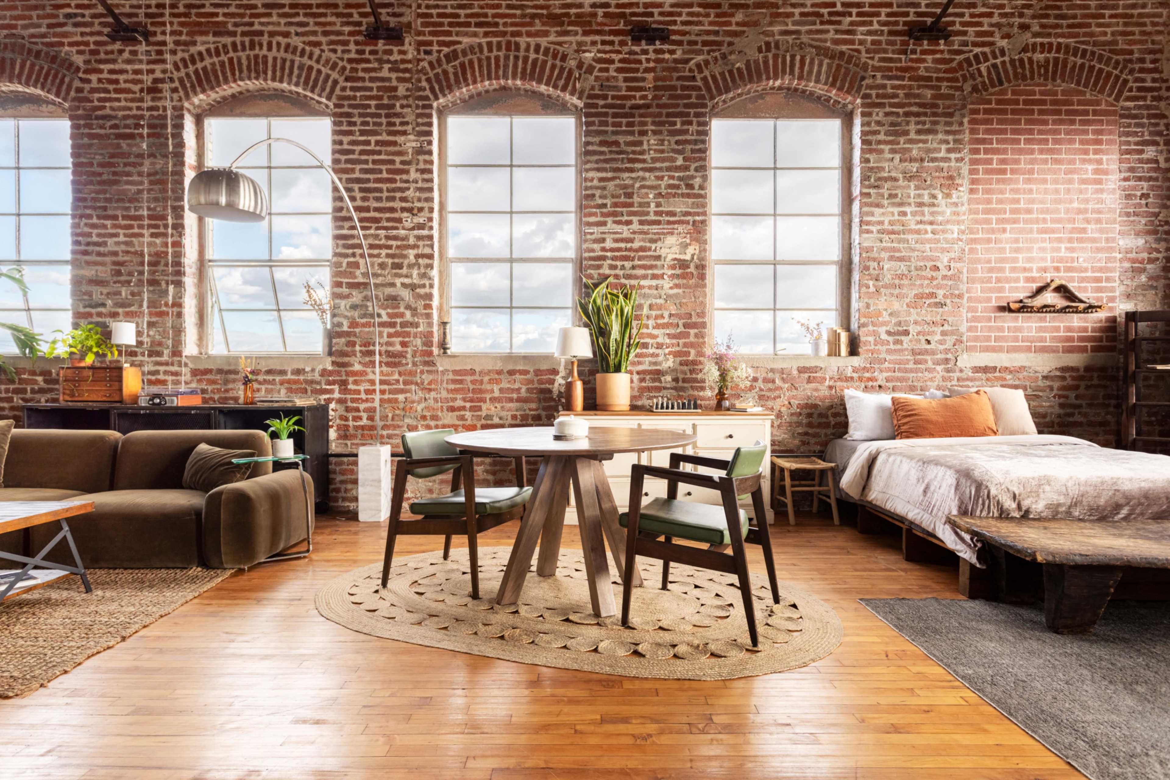 A stylish loft living space featuring a wooden dining table with chairs, a cozy sofa, large windows, and a bed in a room with exposed brick walls.