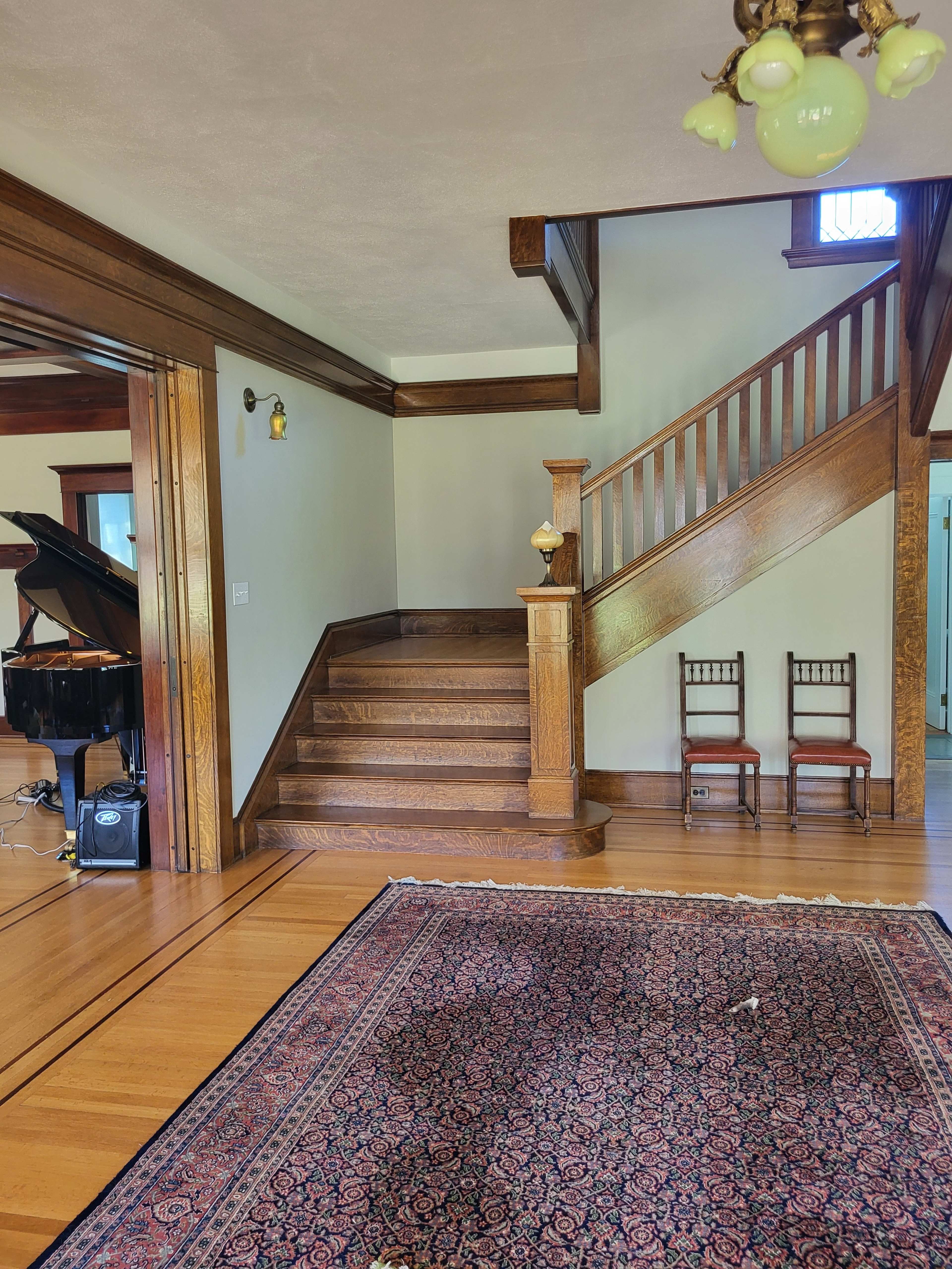 The image shows a spacious interior featuring a staircase, two wooden chairs, a grand piano, and a patterned rug on the hardwood floor.