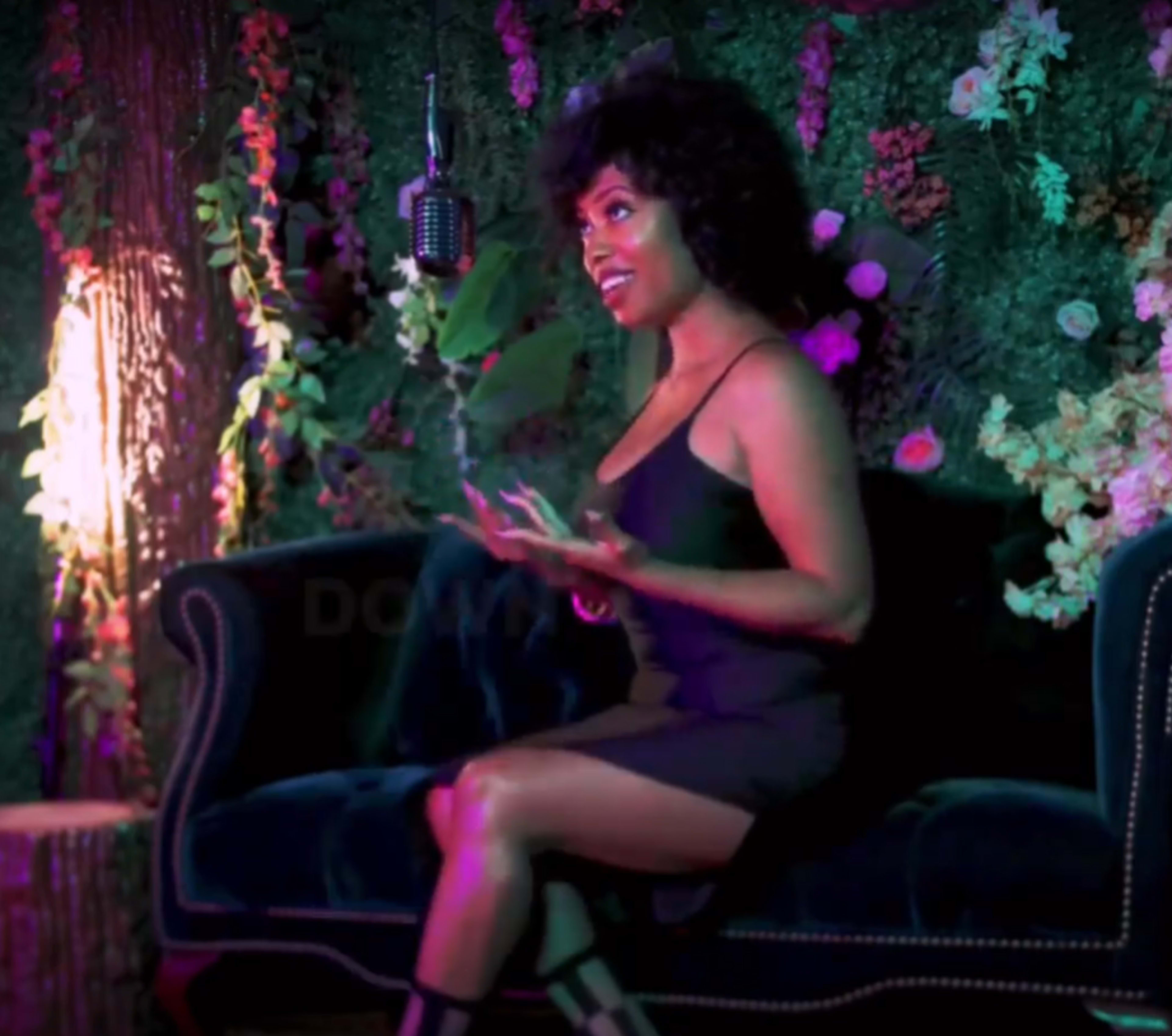 A woman with curly hair is sitting on a dark couch in front of a microphone, surrounded by floral decorations and greenery.