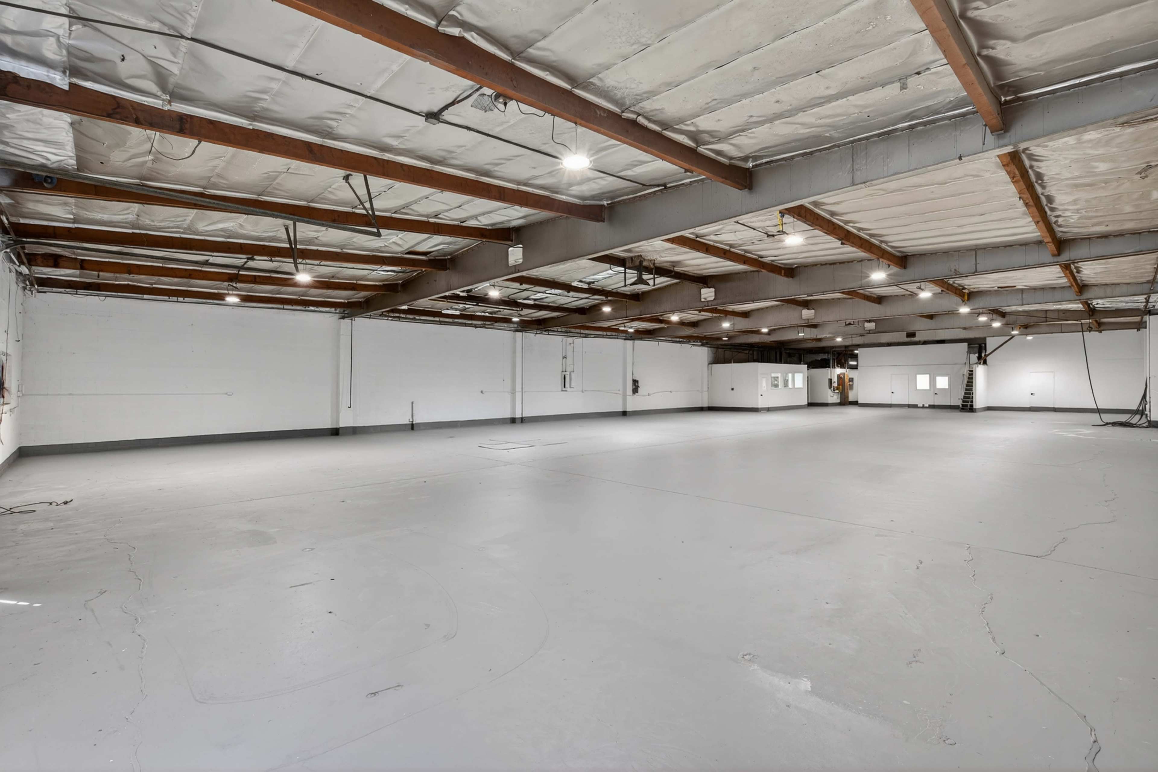 The image shows a large, empty industrial warehouse with concrete flooring and exposed wooden beams in the ceiling.