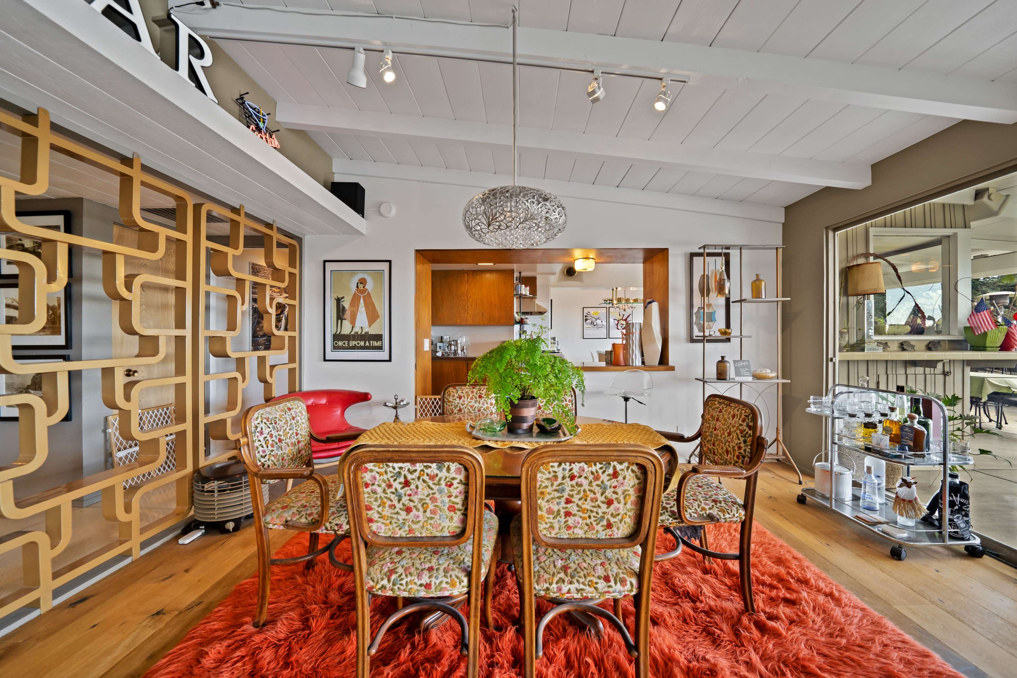 The image shows a dining area with a circular table surrounded by patterned chairs, a large pendant light, and a wooden bar in the background.