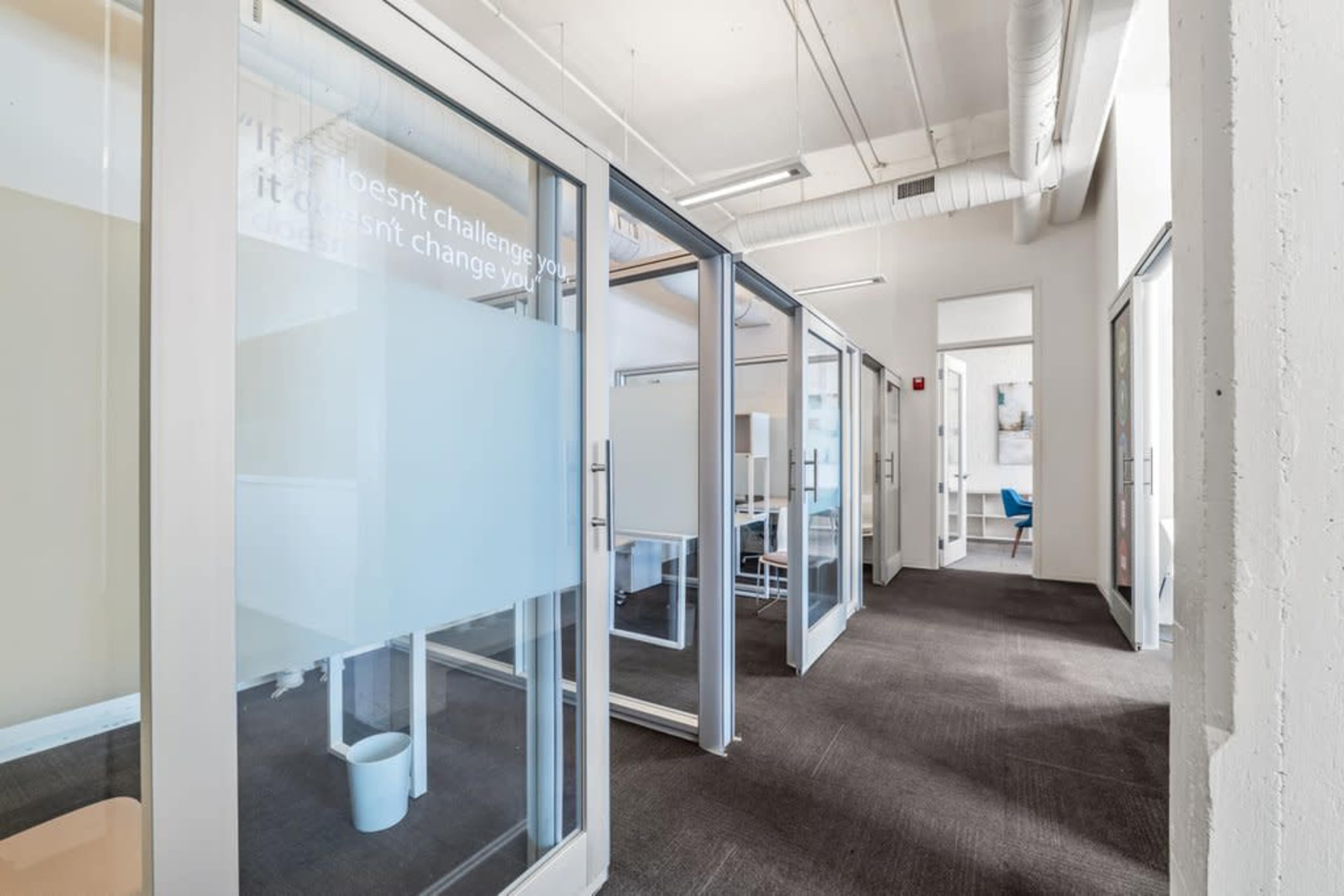 The image shows a modern office corridor with glass-walled meeting rooms on either side and neutral-colored walls.