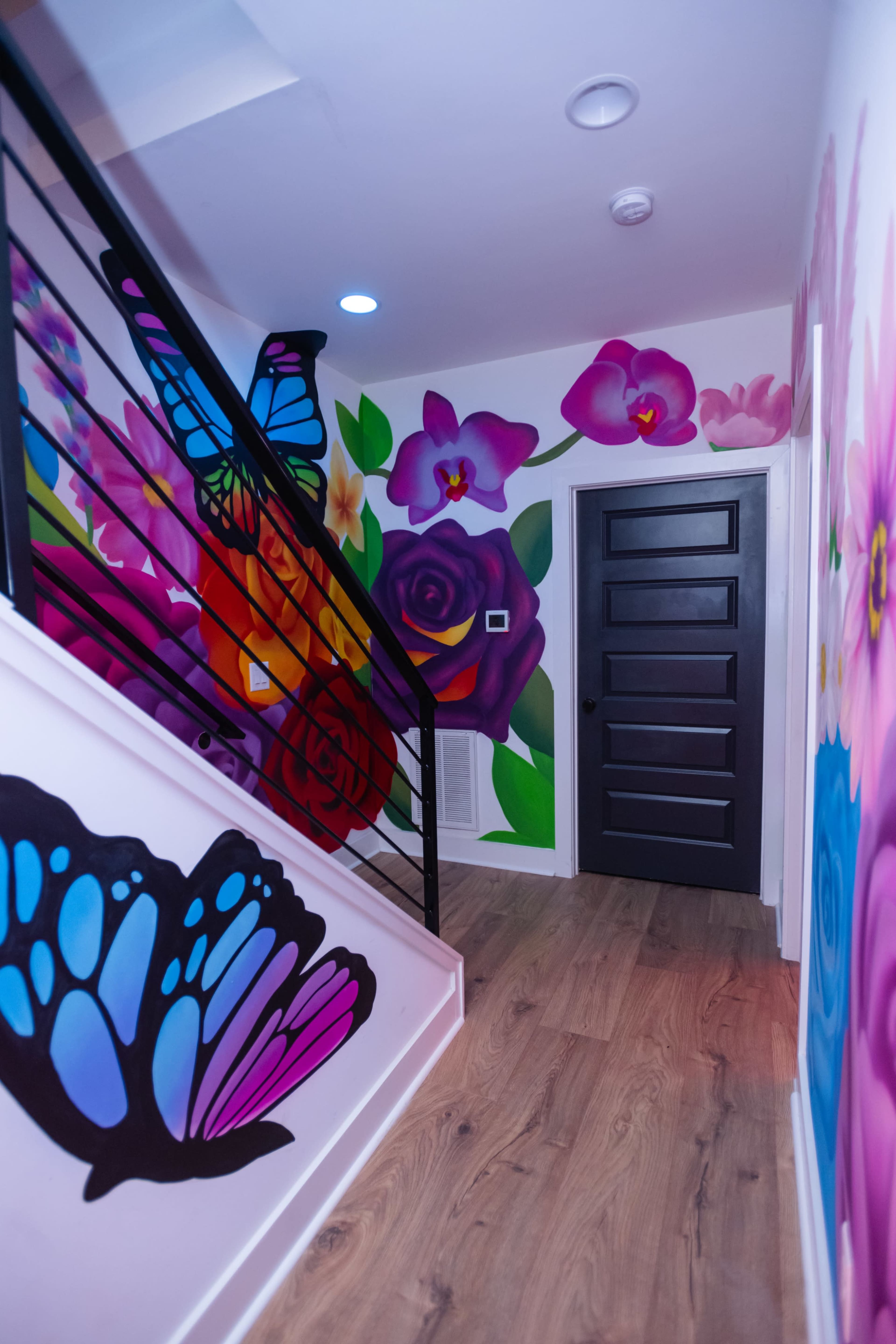 A brightly painted hallway features vibrant murals of butterflies and flowers, leading to a black door at the end of the staircase.