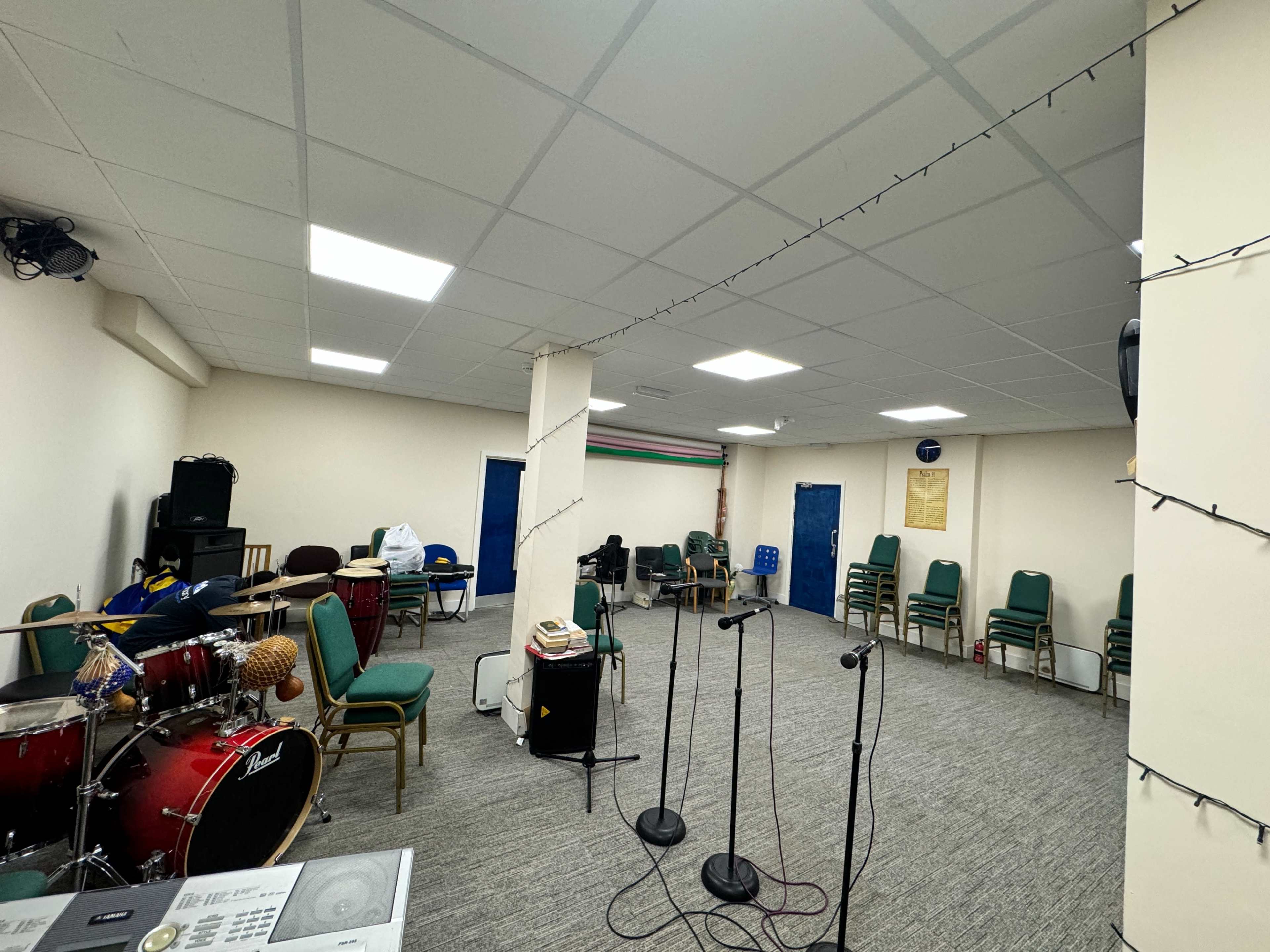 A spacious room set up for music practice, featuring a drum kit, microphones on stands, and stacked chairs against the walls.
