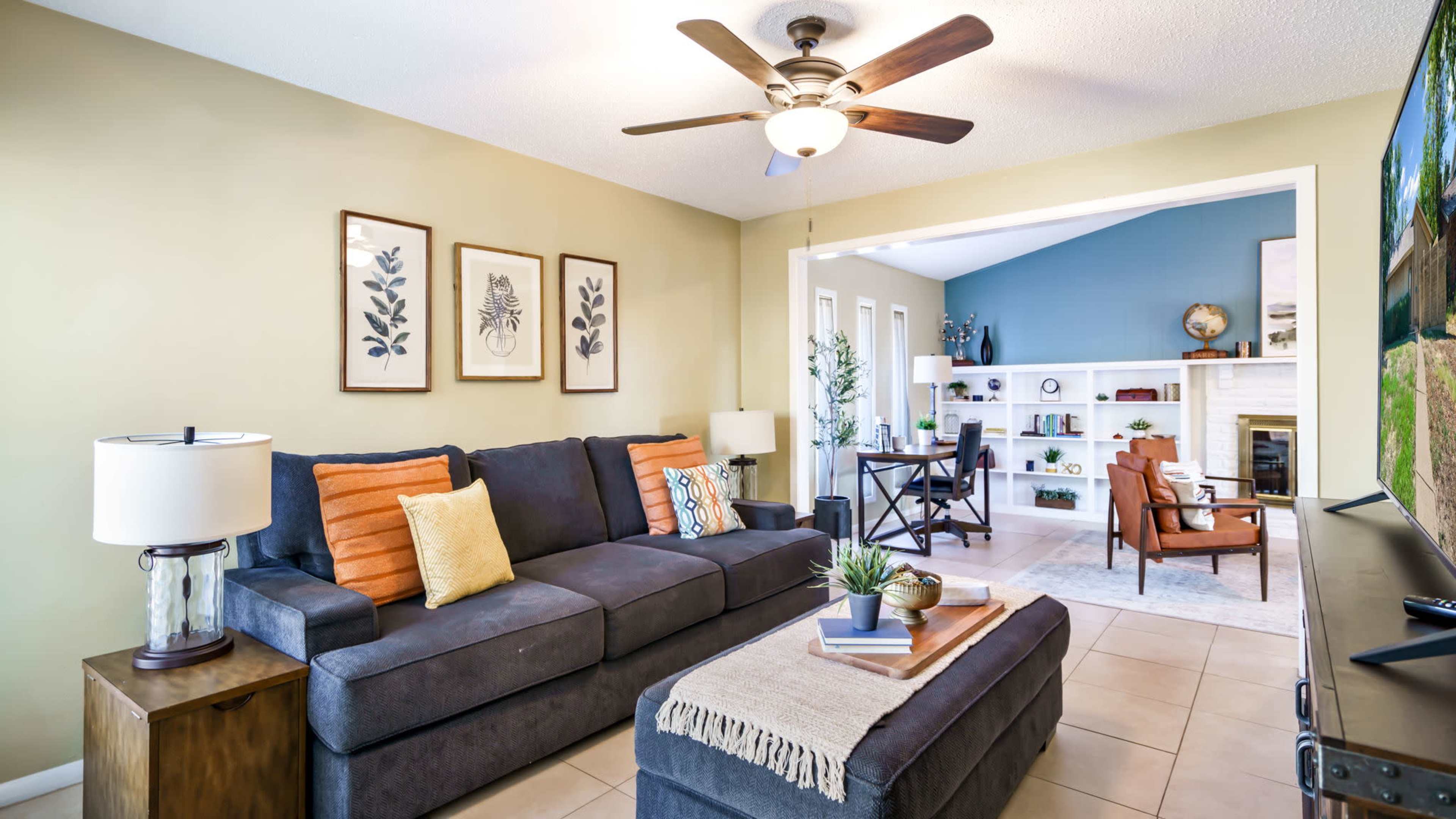 A cozy living room features a dark sofa with colorful pillows, a coffee table, and a fan above, with an open space leading to a brightly decorated room.