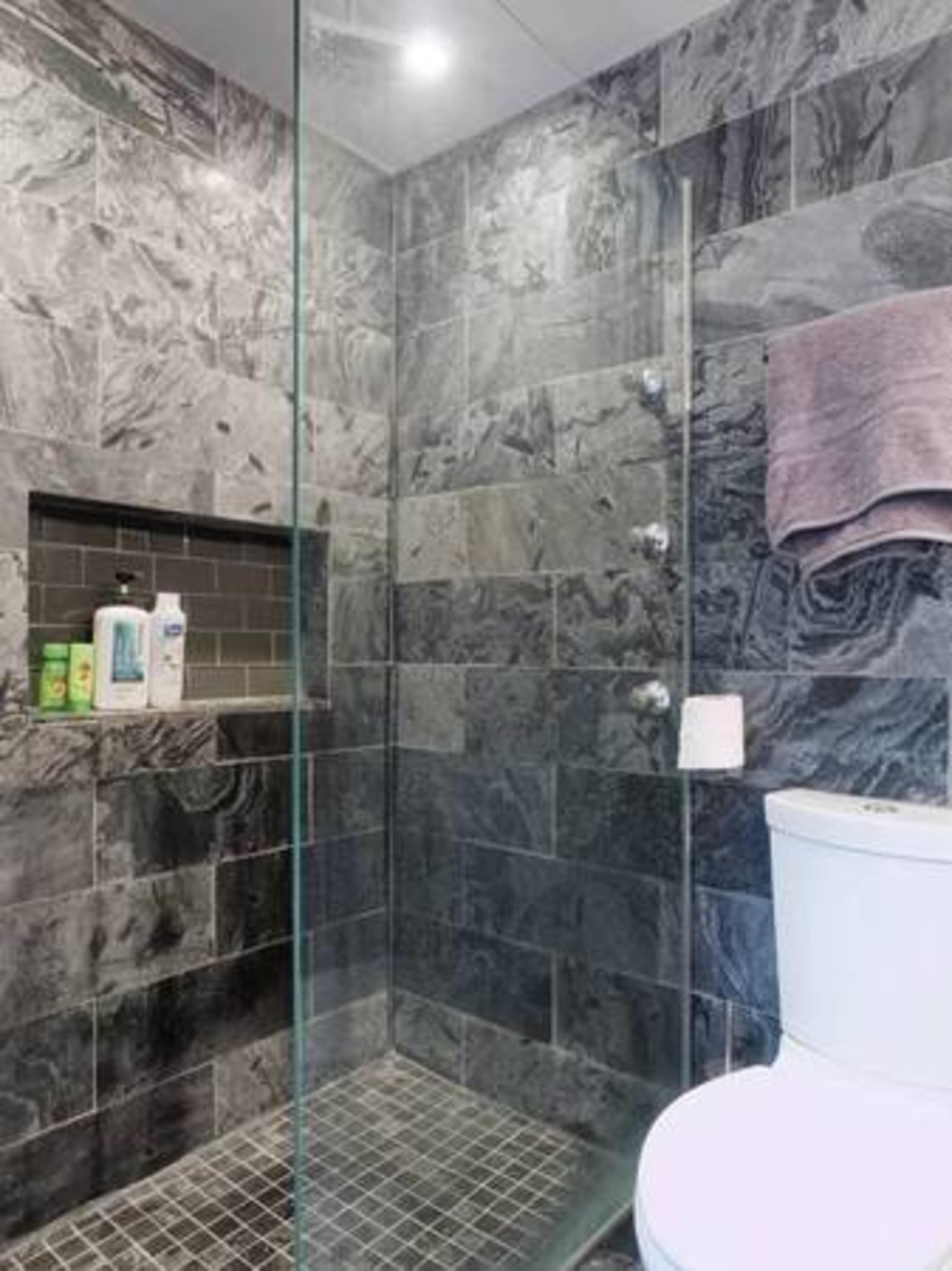 A modern shower enclosed in glass, featuring dark stone walls, a tiled floor, and a small shelf for toiletries.