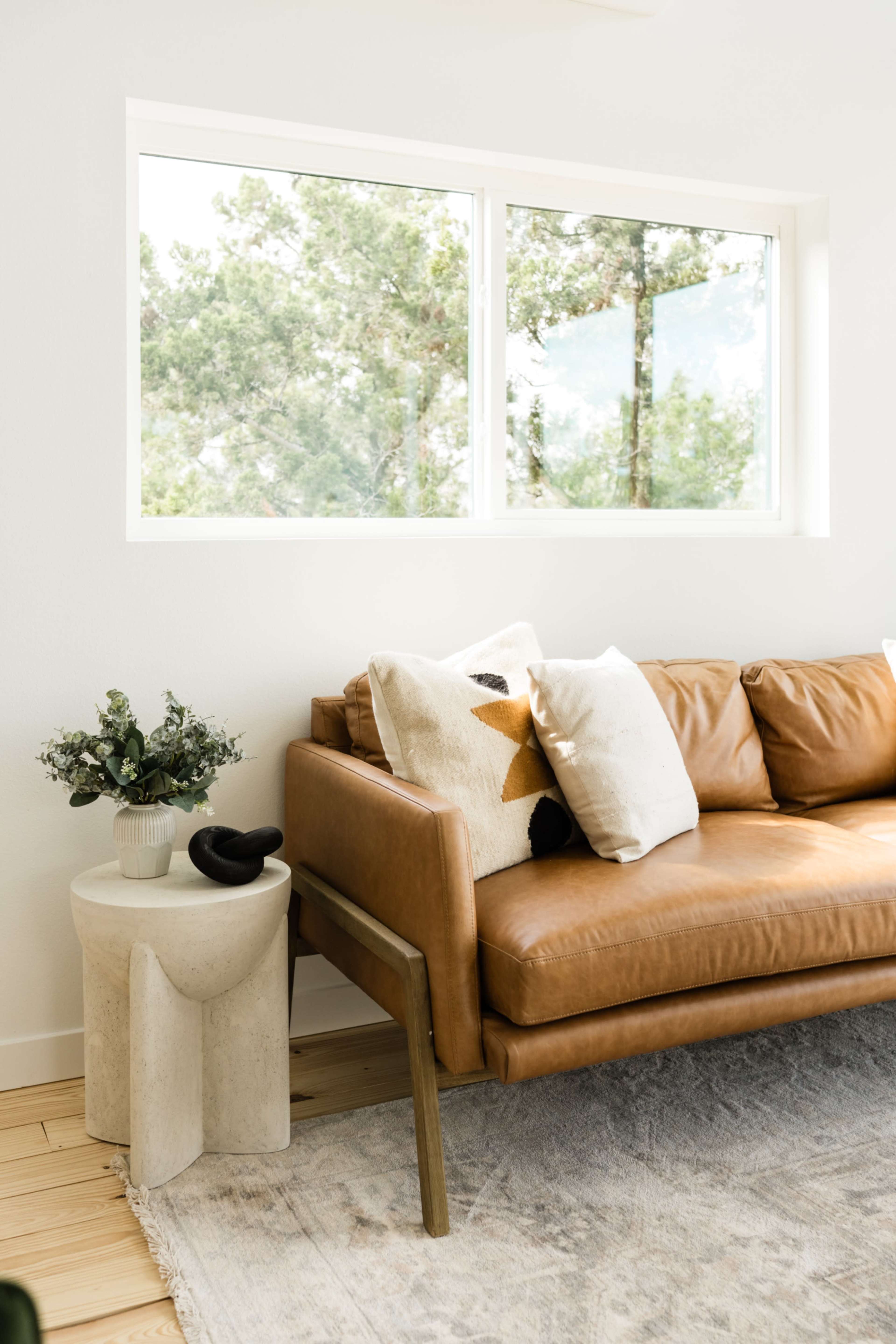 A brown leather sofa with patterned cushions is positioned next to a light-colored side table, beside a large window offering a view of trees outside.