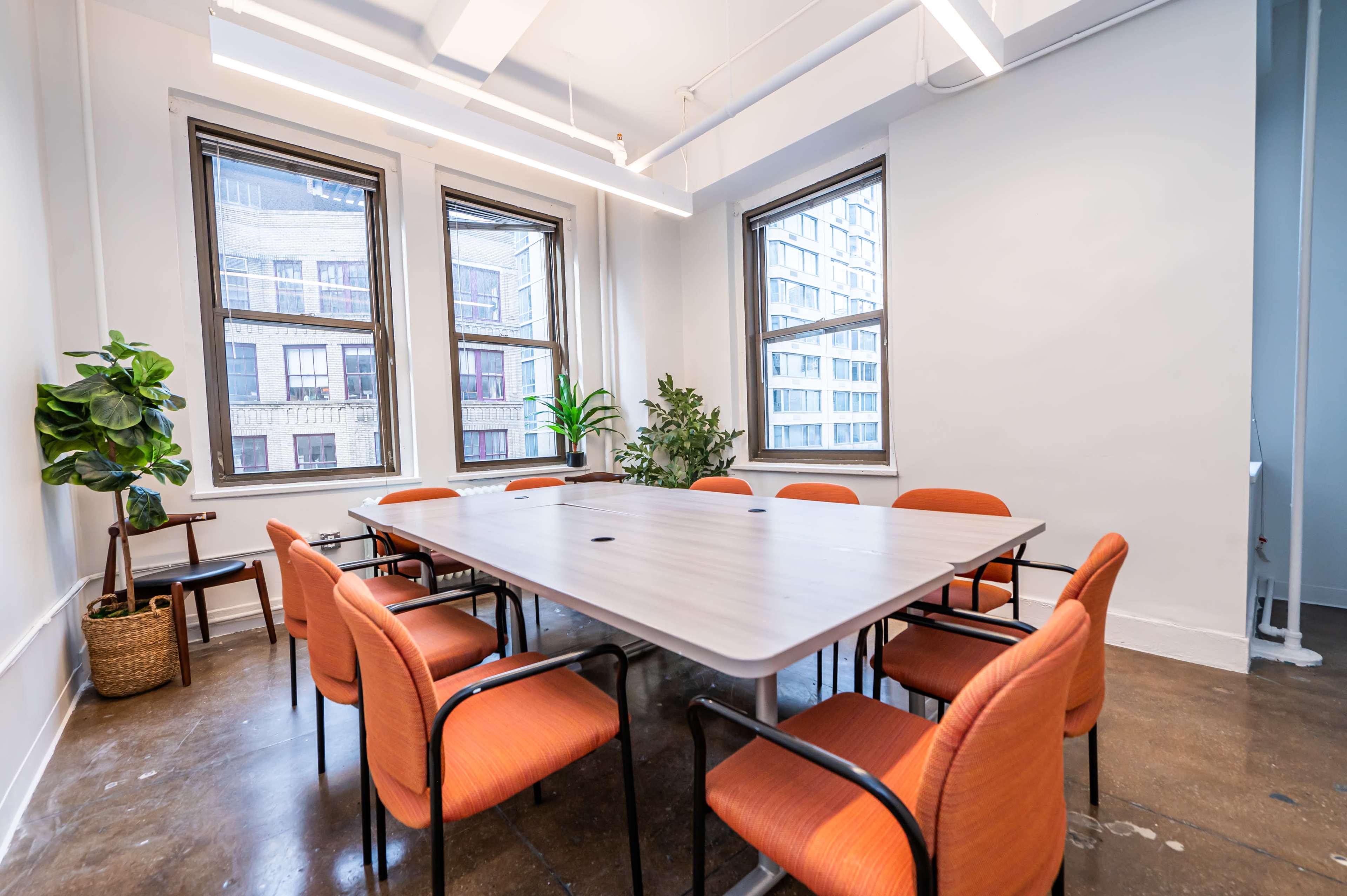A conference room features a large table surrounded by orange chairs and has windows with city views.