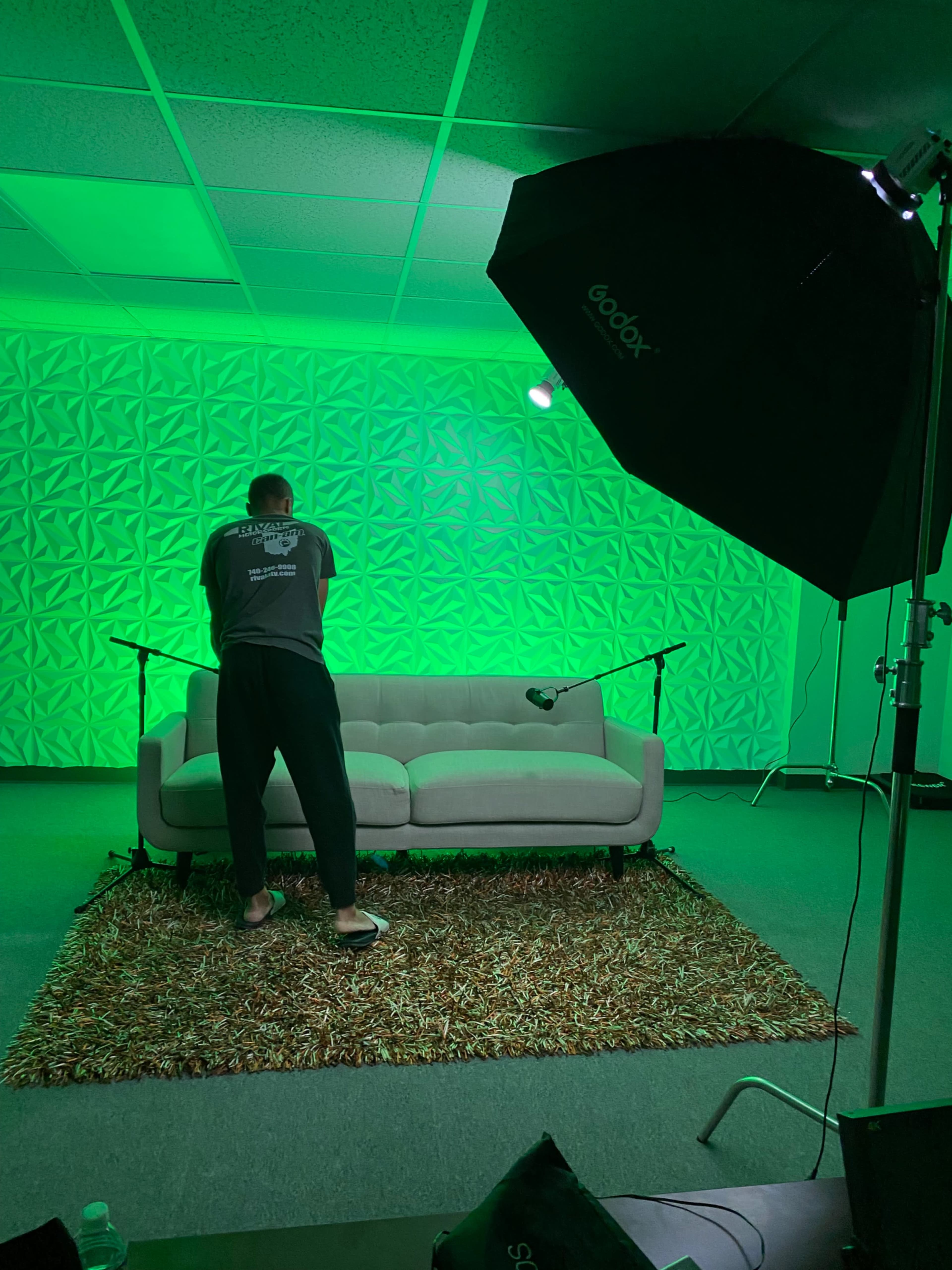 A person stands in front of a green-lit wall, adjusting equipment near a gray couch on a textured rug.