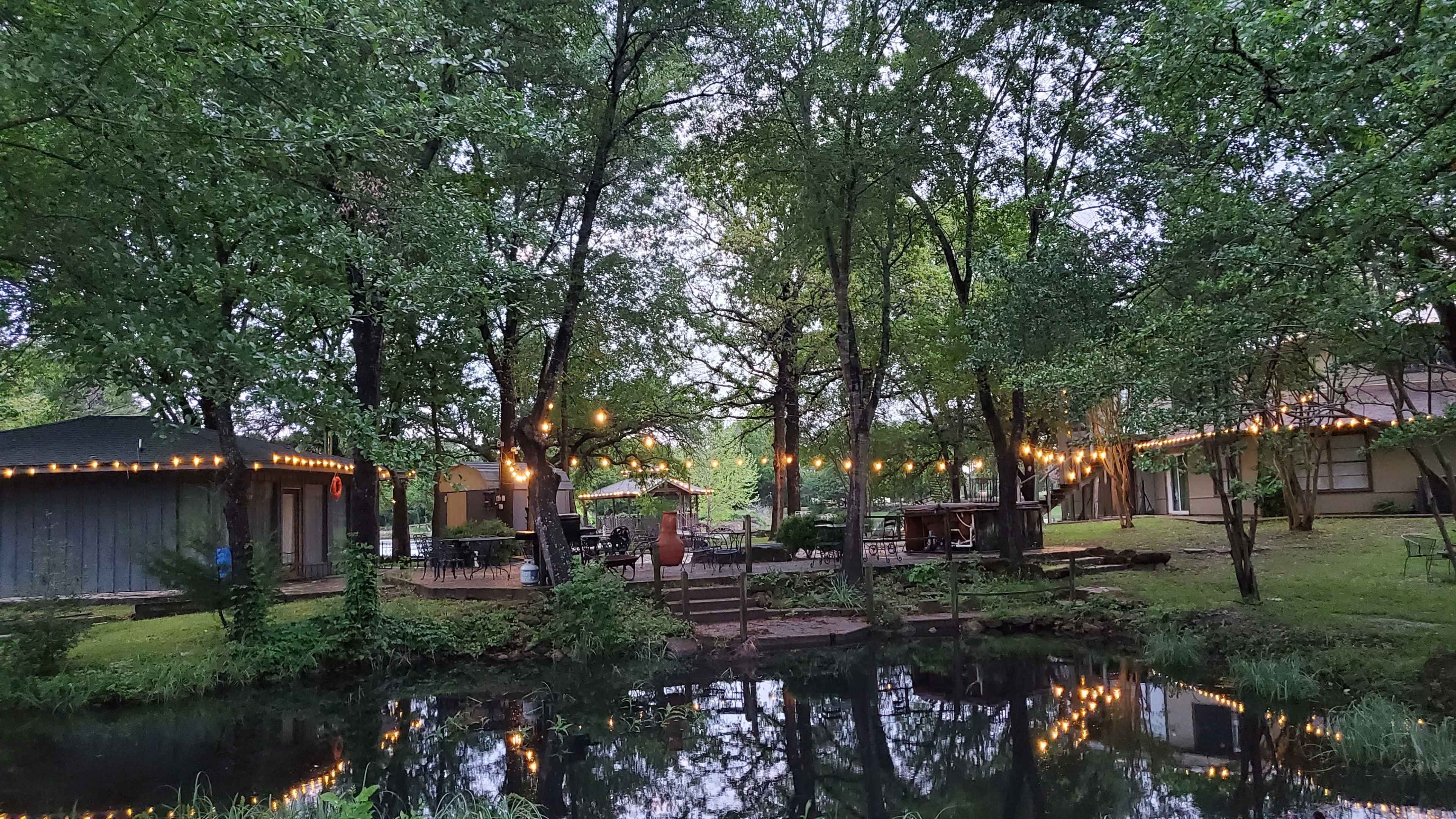 A serene outdoor space features a pond surrounded by trees, with string lights hanging overhead and several structures nearby.