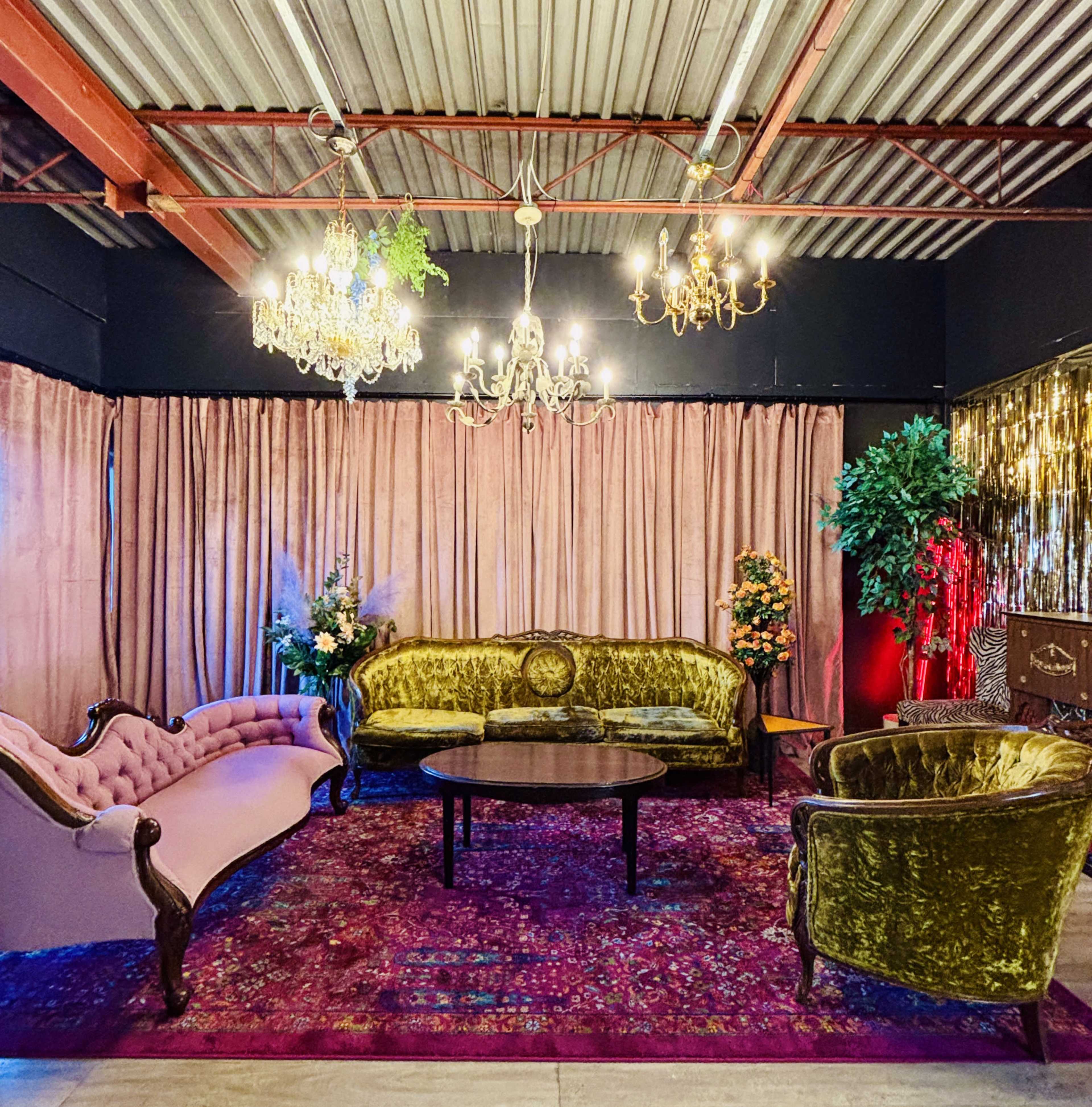 The image shows a colorful lounge area featuring vintage sofas, a round table, and decorative plants, all set against a backdrop of velvet curtains and chandeliers.