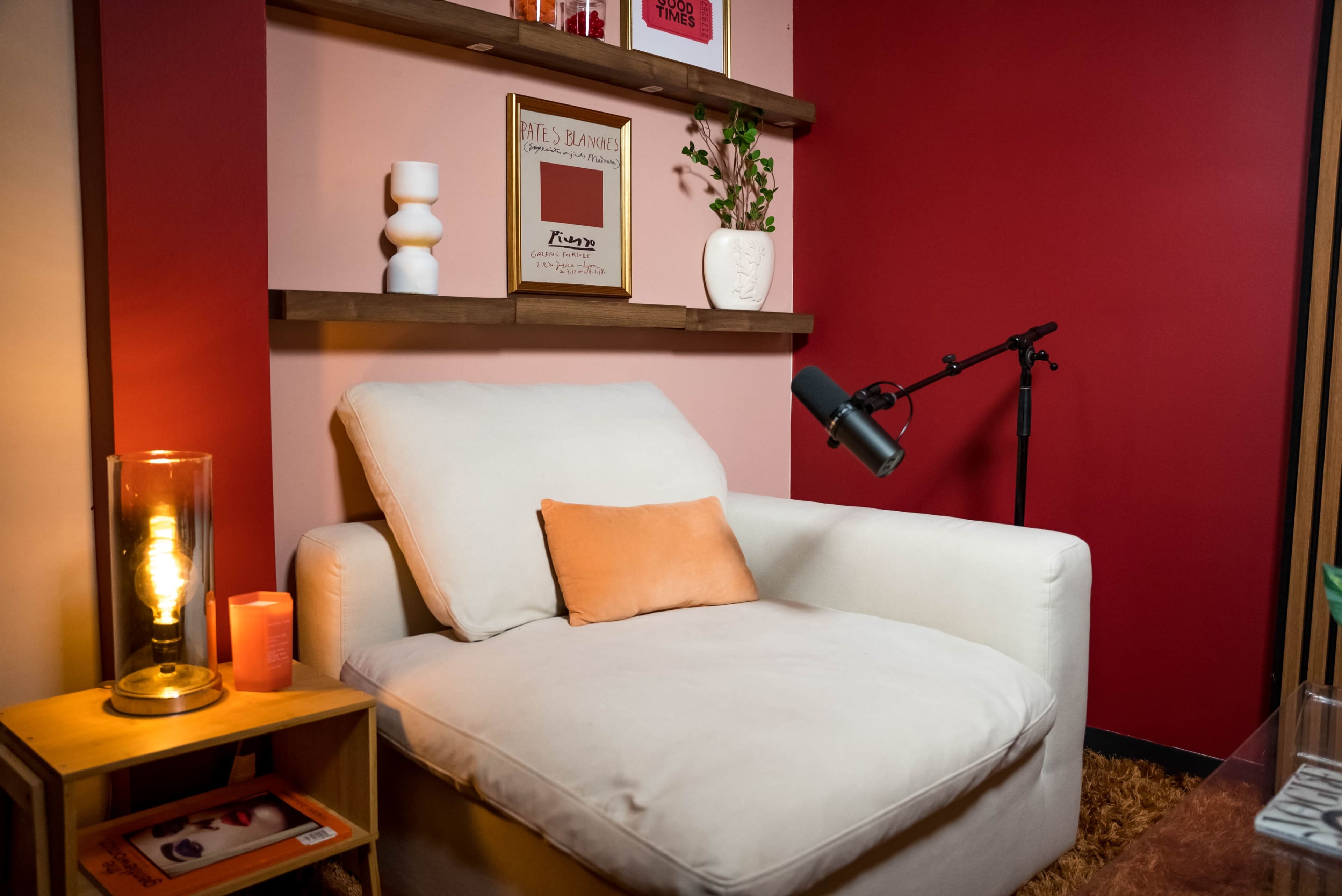 A cream-colored armchair is positioned next to a wooden side table with a lamp and decorative items against a backdrop of red walls and a shelf displaying a small plant and framed art.