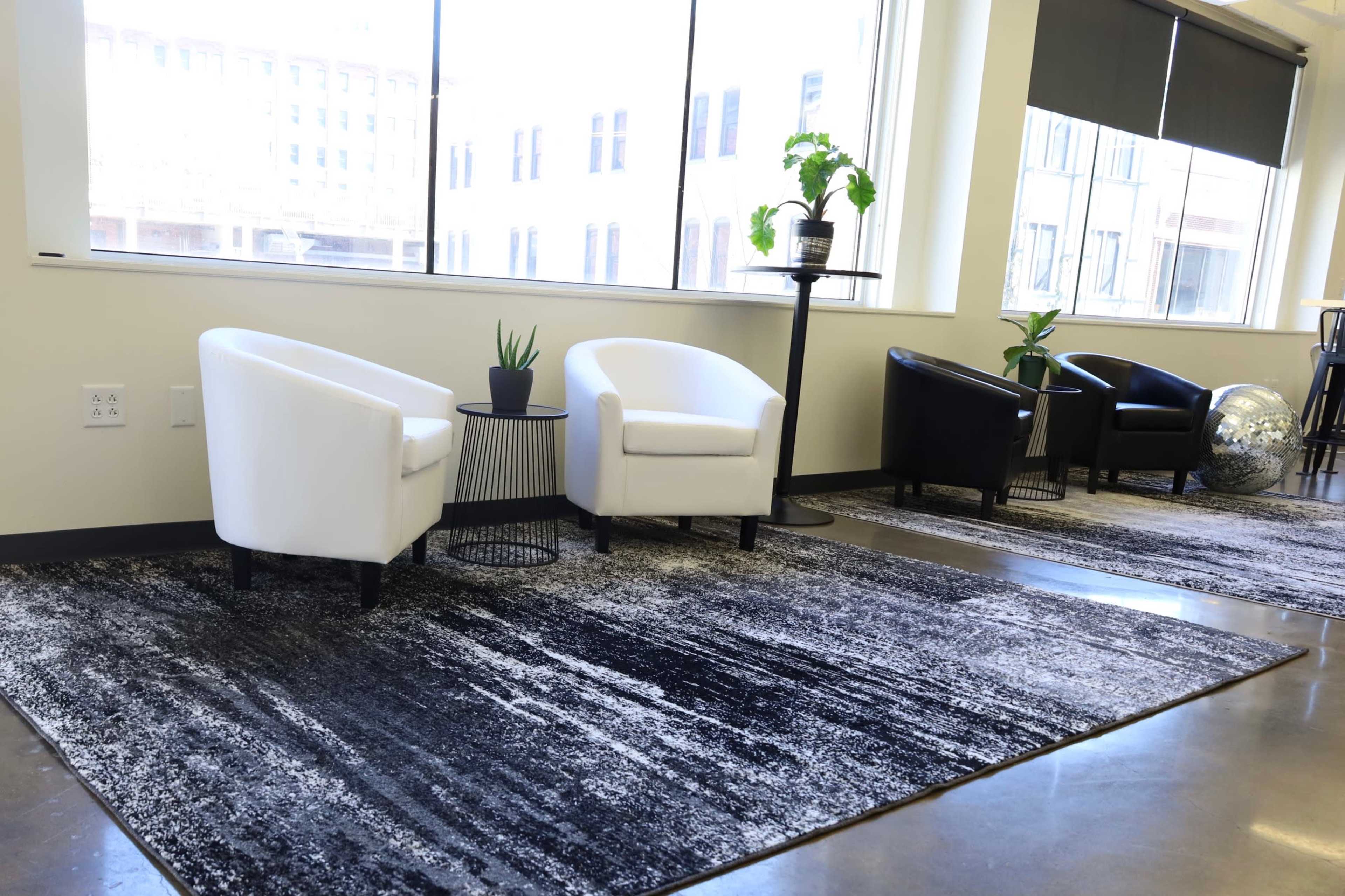 A modern seating area with two white armchairs and one black armchair, accompanied by small tables and greenery, set against large windows.