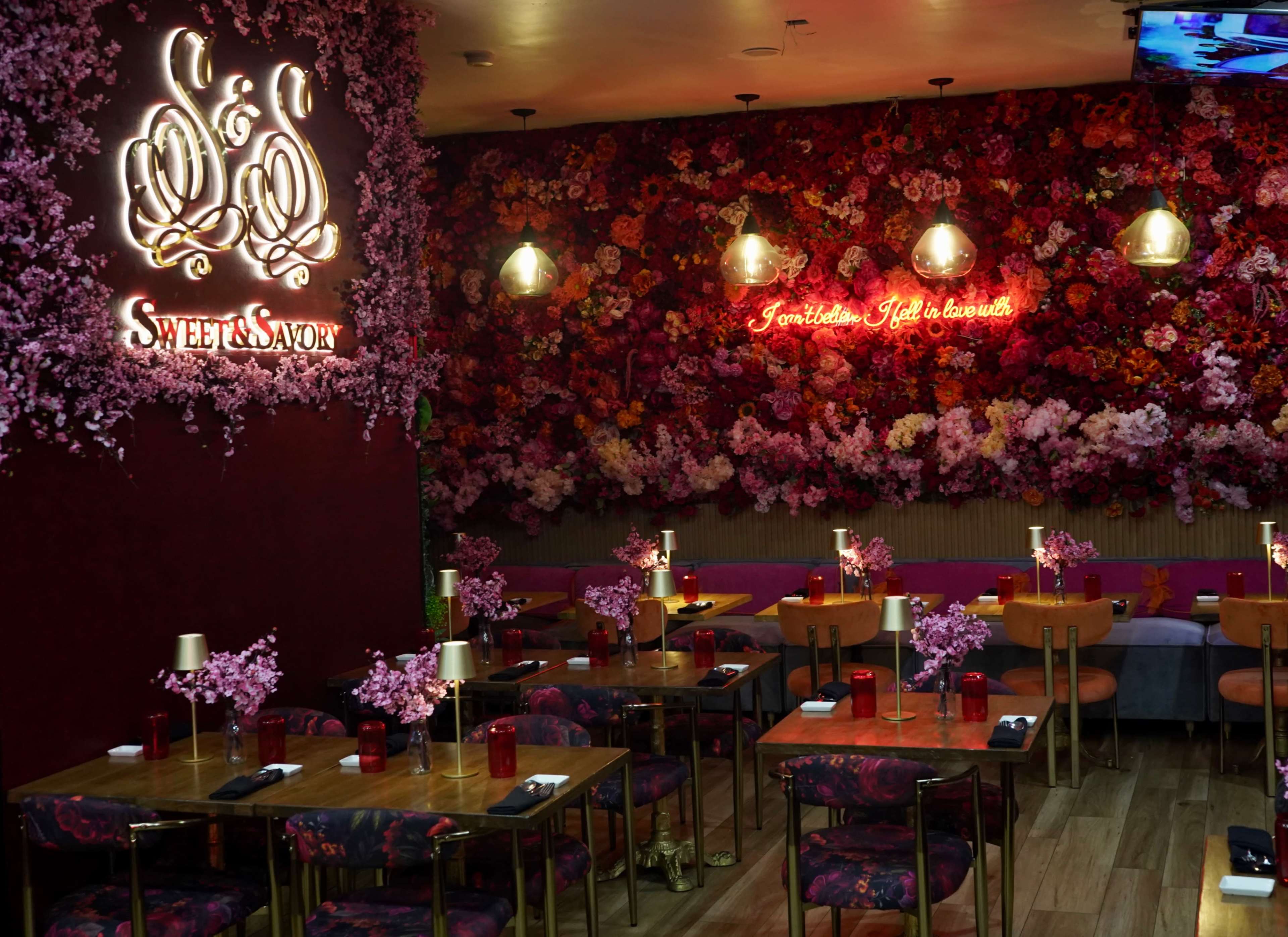 The interior of a restaurant features wooden tables with floral centerpieces, surrounded by a wall covered in vibrant artificial flowers and illuminated signs.
