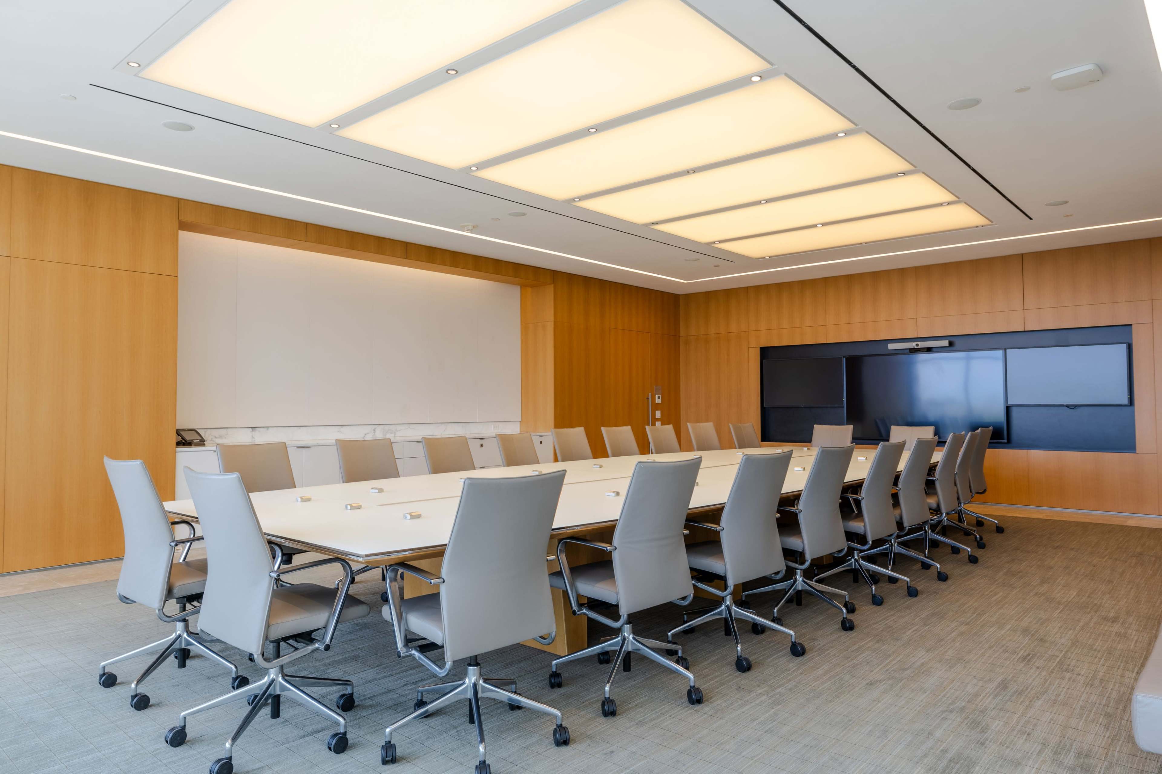 The image shows a modern conference room with a large table surrounded by ergonomic chairs, illuminated by a ceiling light panel.