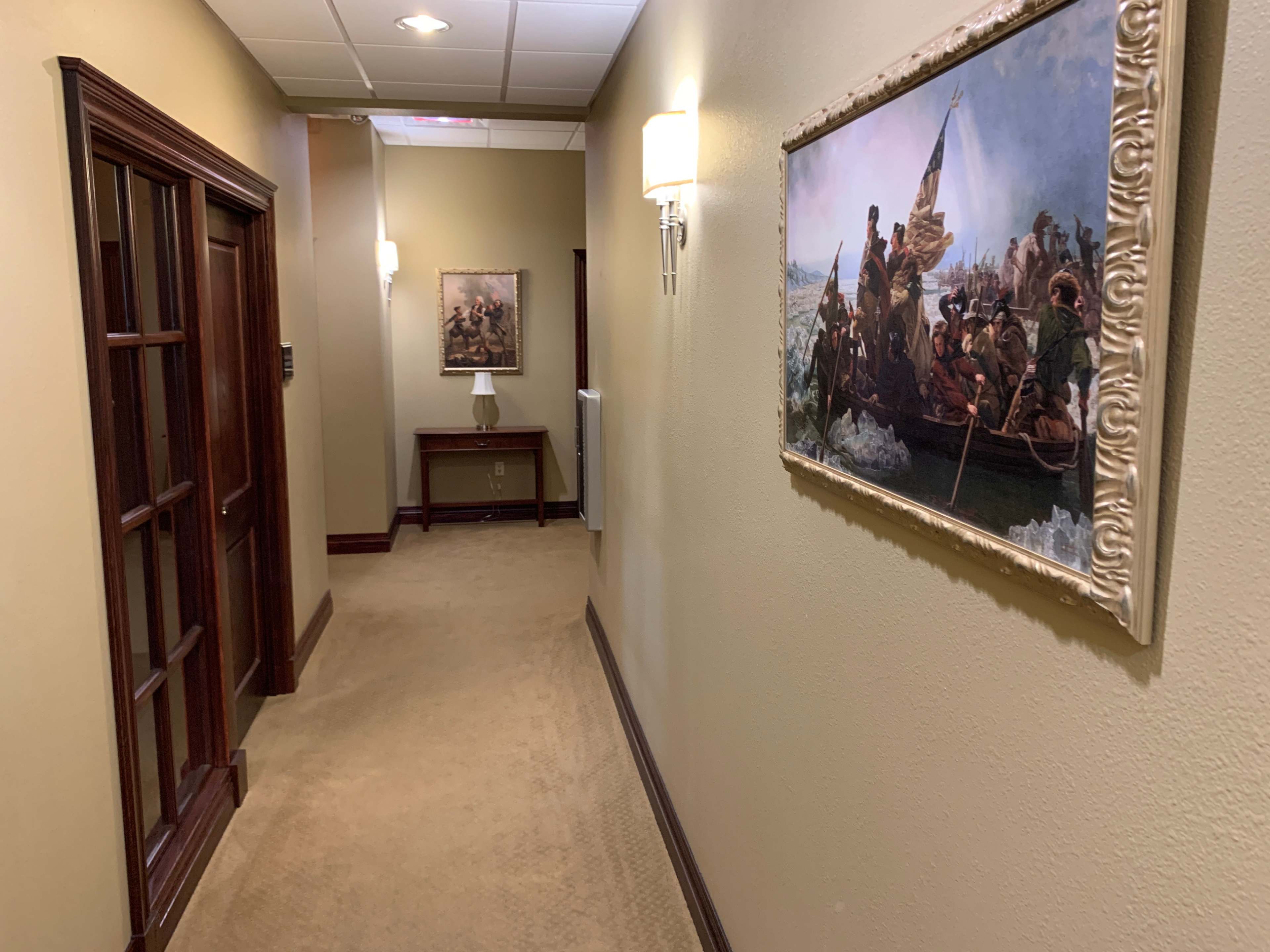 A well-lit hallway features two framed paintings on the walls and a table with a lamp at the end.