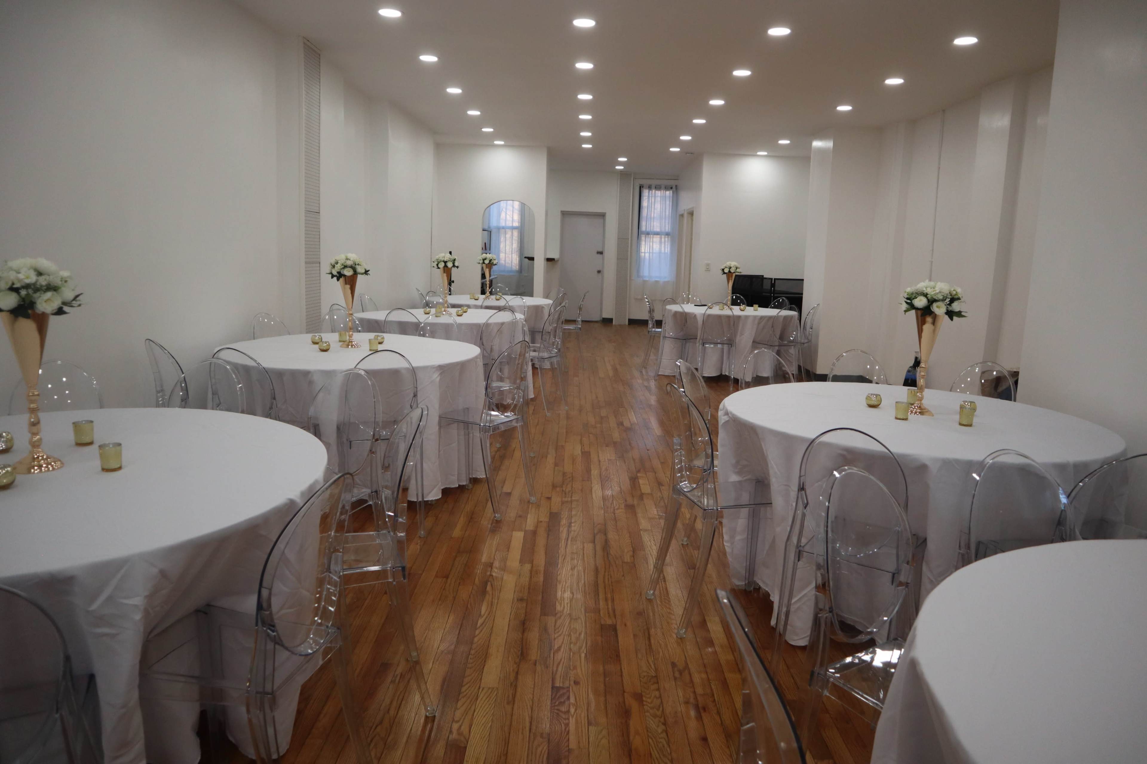 An empty event space with several round tables covered in white tablecloths, surrounded by transparent chairs, and adorned with small floral centerpieces.