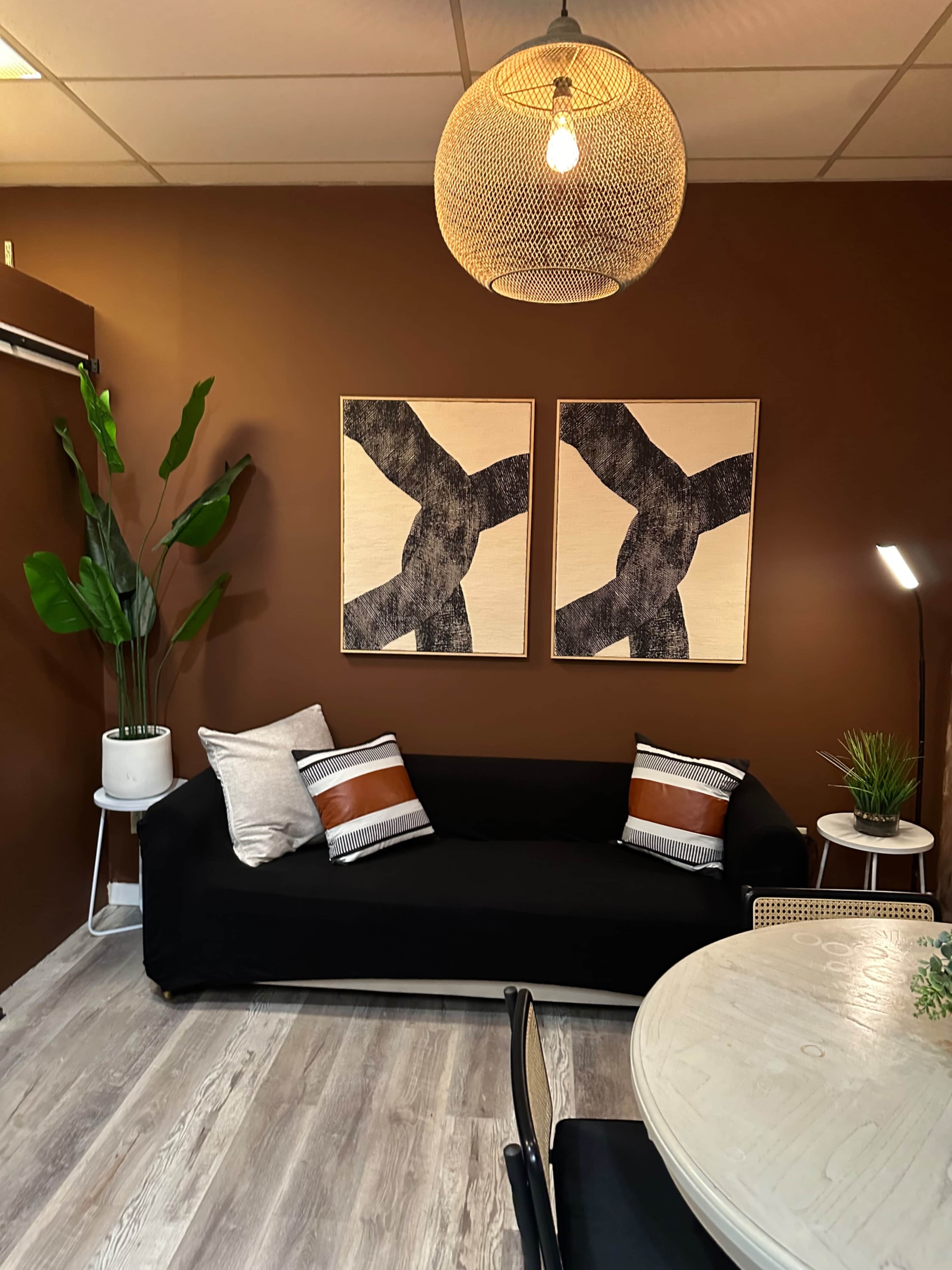 The image shows a modern room featuring a black couch with decorative pillows, two abstract wall artworks, a potted plant, a round table, and a pendant light against a brown wall.