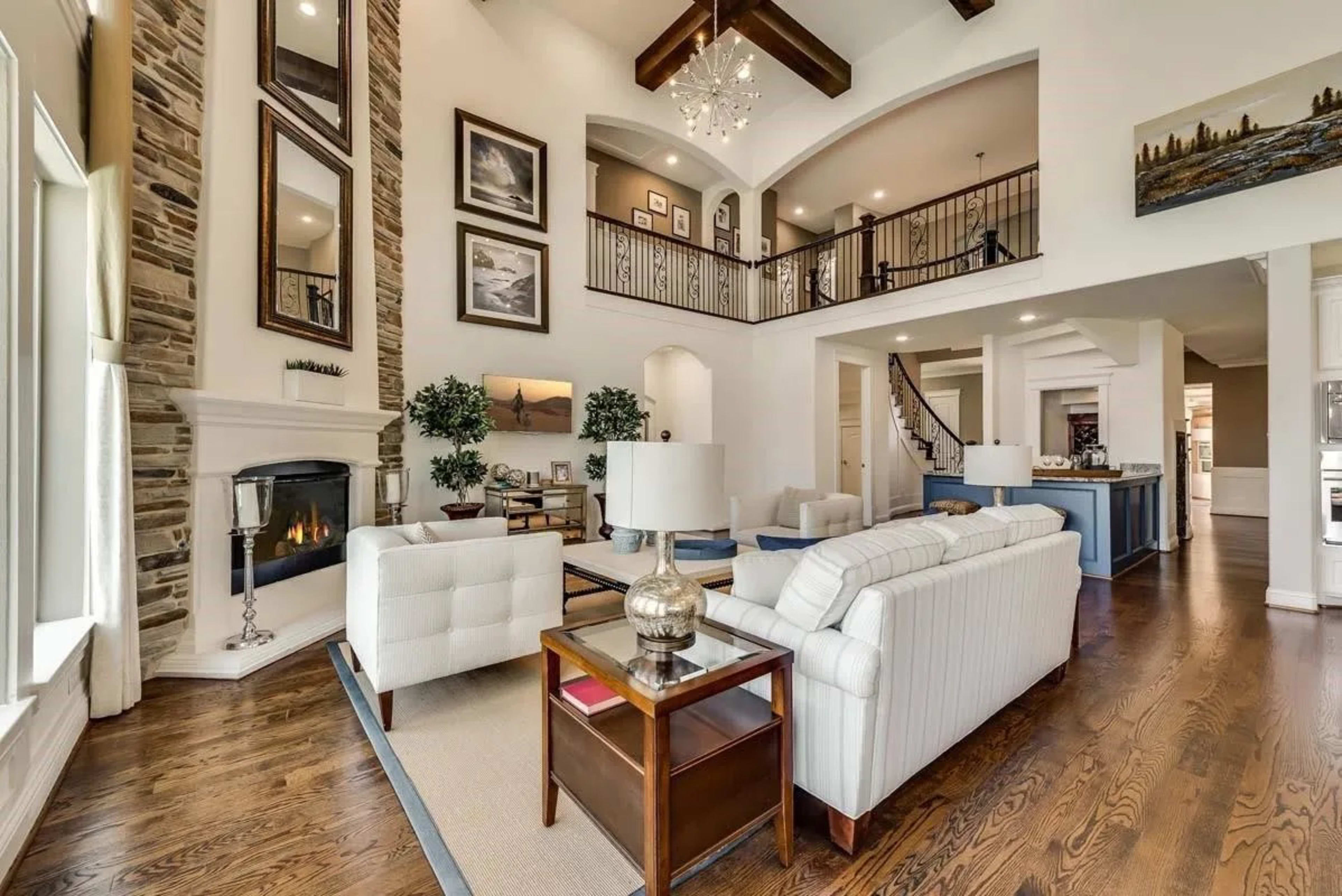 The image depicts a spacious living area with high ceilings, a fireplace, and a staircase leading to an upper level, featuring modern furniture and decorative elements.