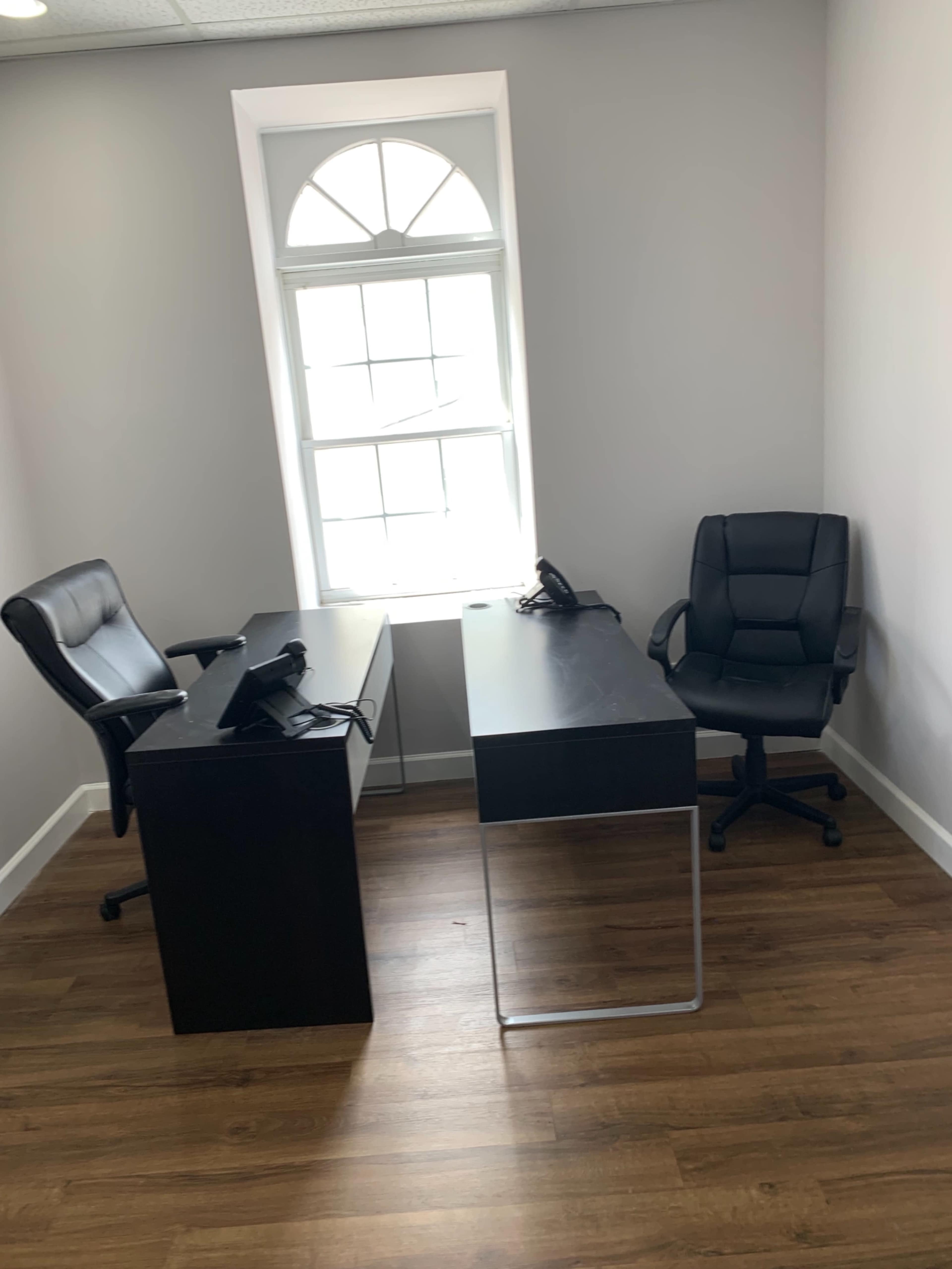 The image shows a small office space with two desks, one black and one sleek, along with two chairs and a window letting in natural light.