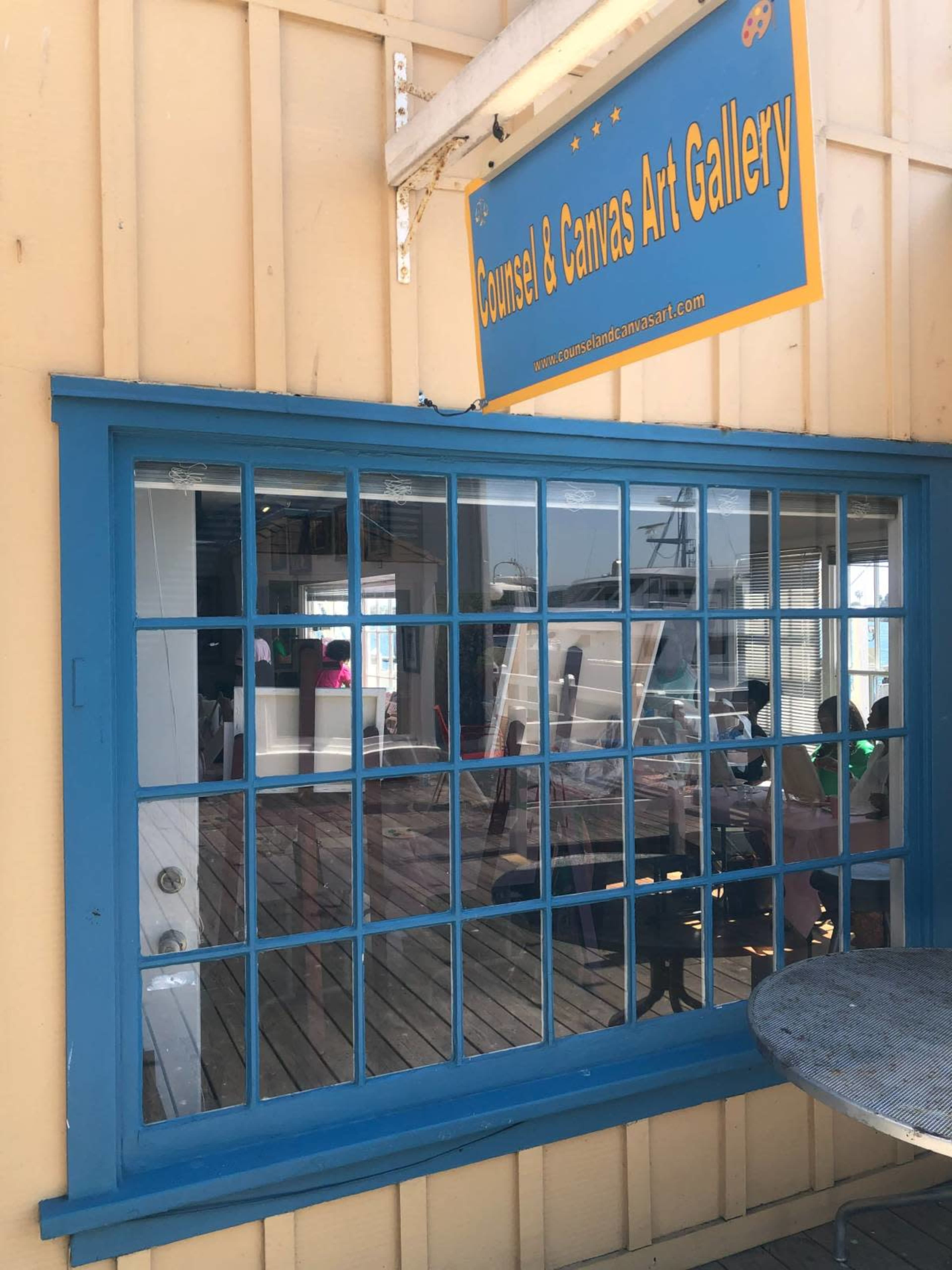 The image shows a blue-framed window reflecting a wooden deck and the interior of an art gallery, with a sign above that reads "Counsel & Canvas Art Gallery."