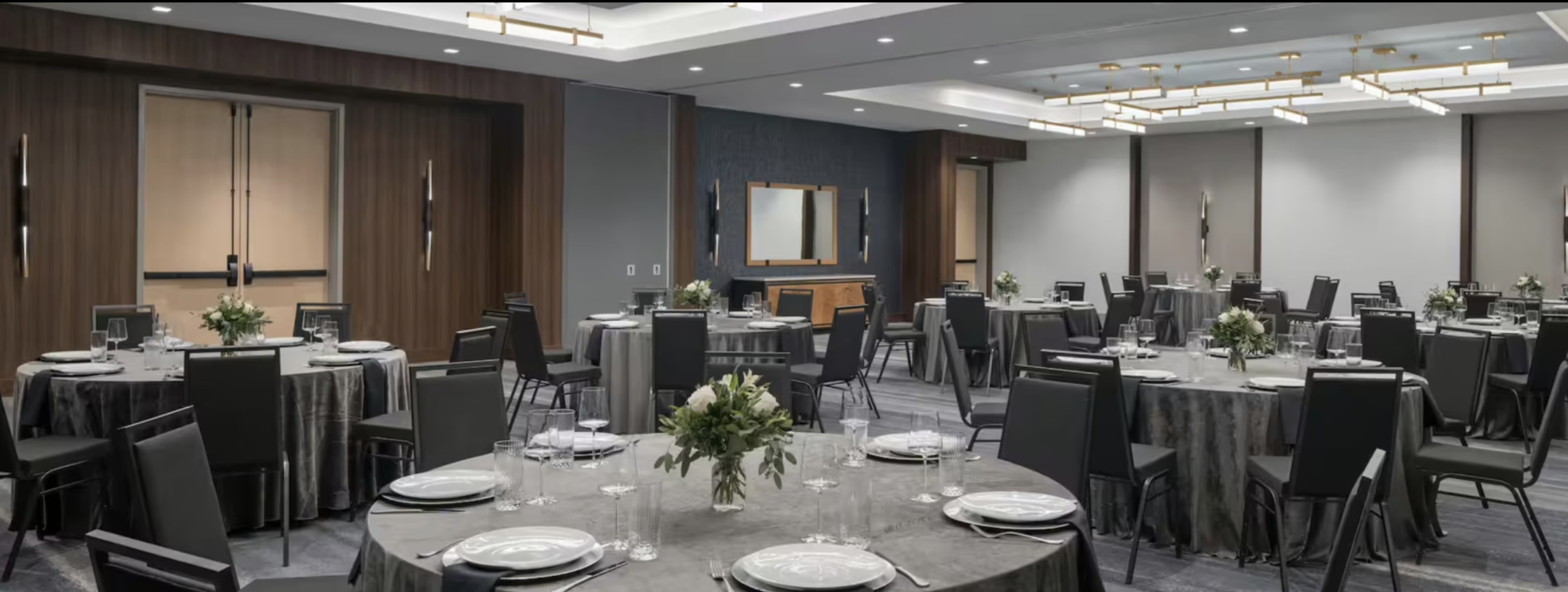 The image shows a banquet hall set up with round tables adorned with gray tablecloths, each featuring centerpieces of flowers, and black chairs arranged around them.