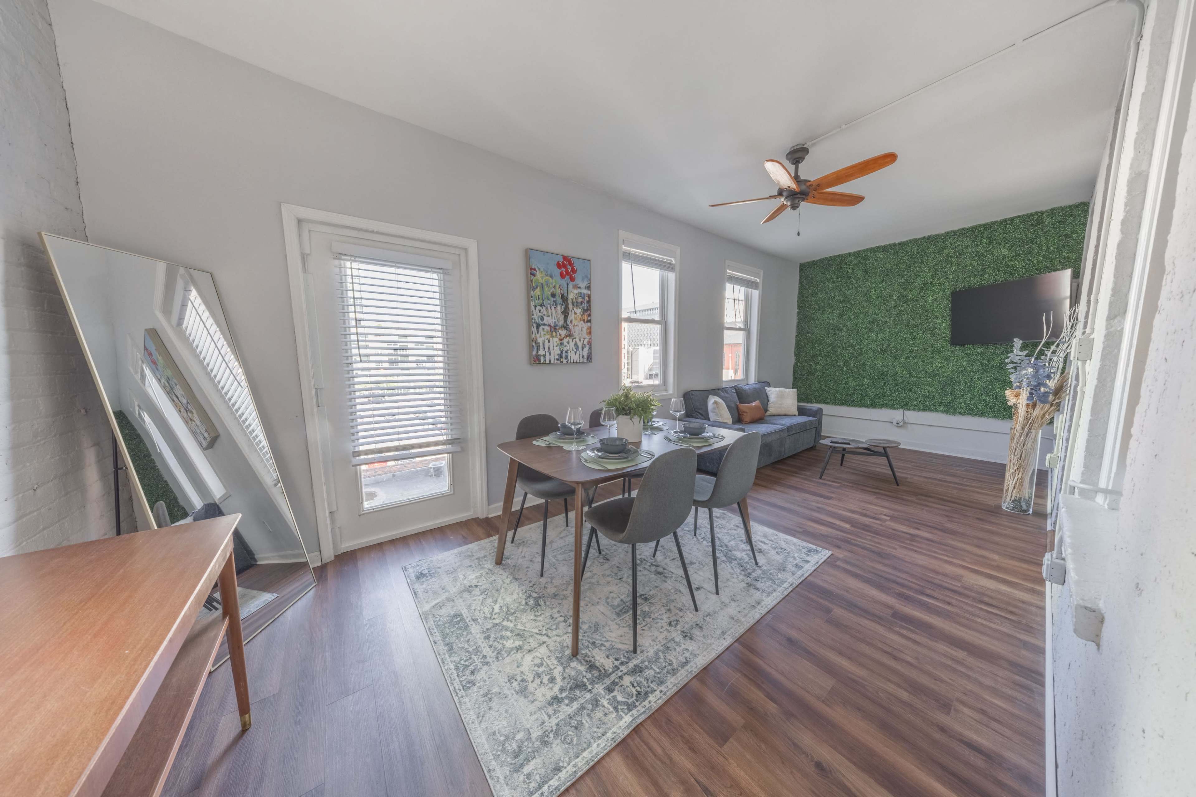 The image shows a modern living space featuring a dining area with a table set for four, a green wall, and natural light streaming through windows.