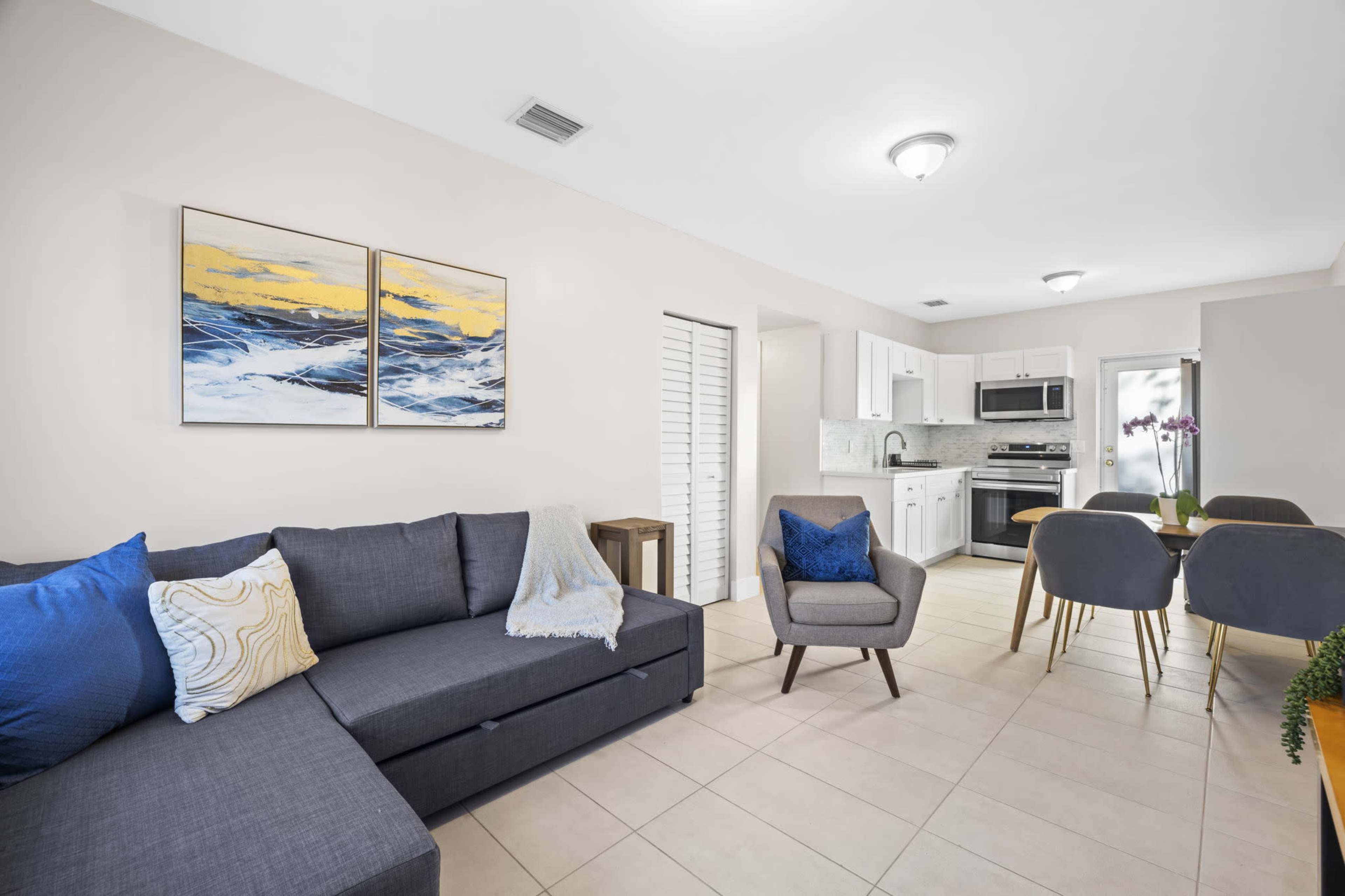 The image shows a modern living area with a gray sectional sofa, a small dining table, and a kitchen in the background, featuring light-colored tiles and a sliding door.