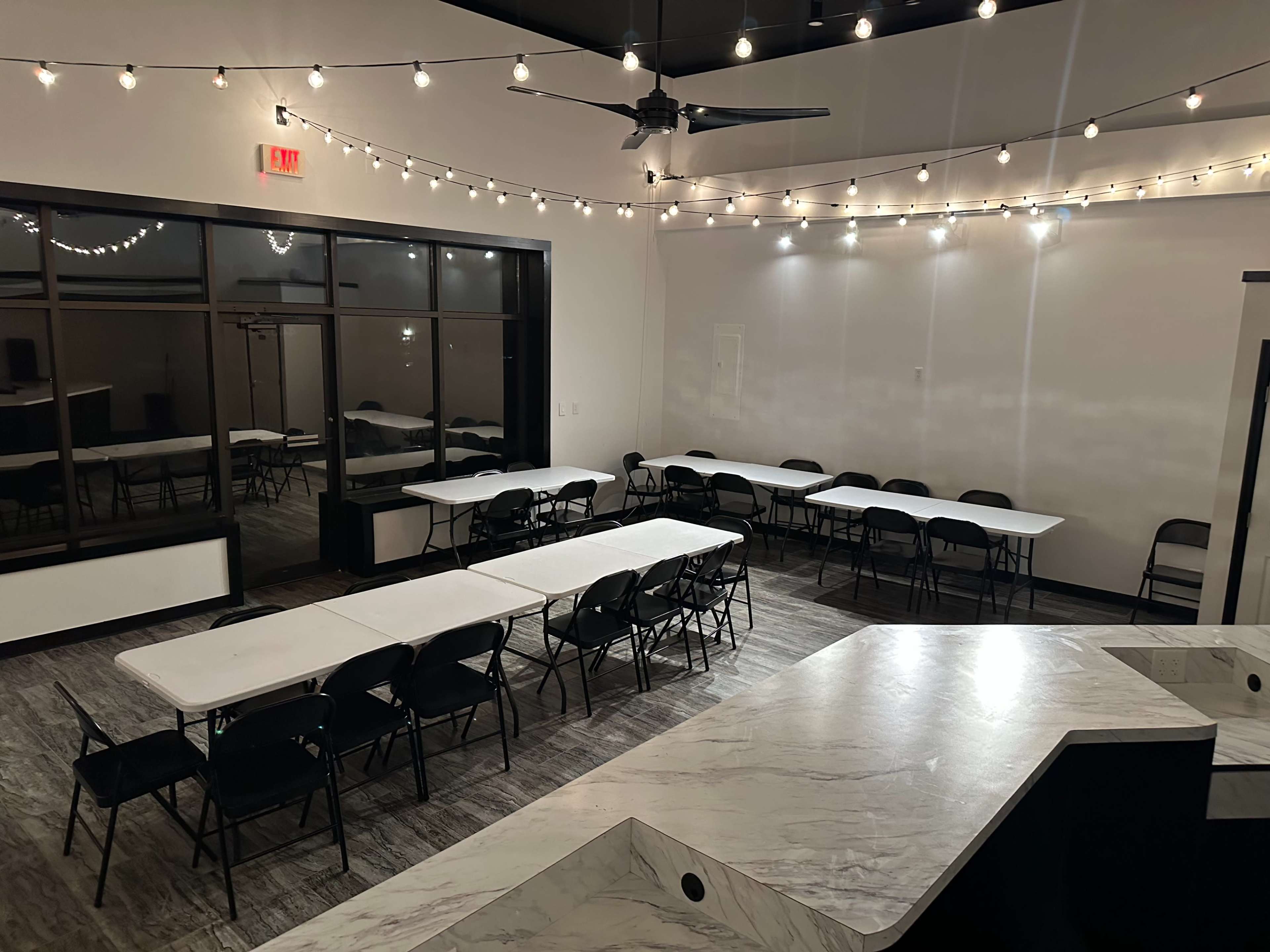 The image shows a modern event space with multiple rectangular tables and chairs arranged under string lights, creating a bright and open atmosphere.