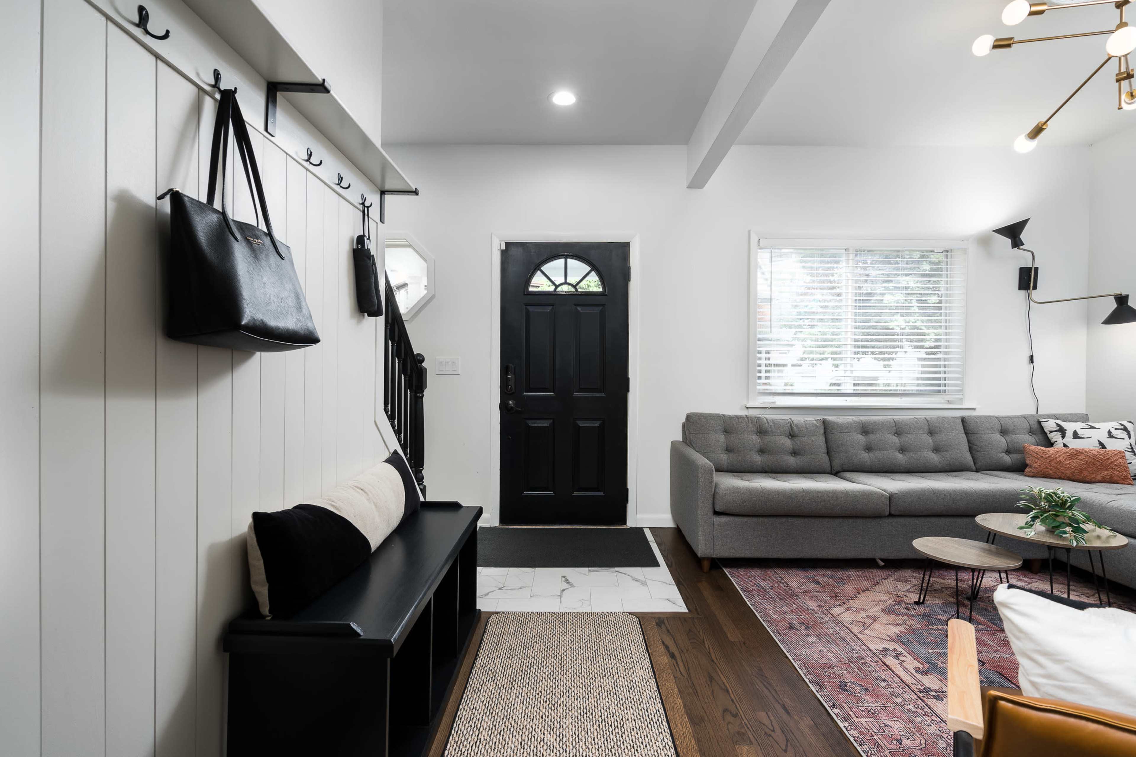 A modern entryway featuring a black door, a gray sofa, a coat rack with bags, and a small coffee table on a rug.
