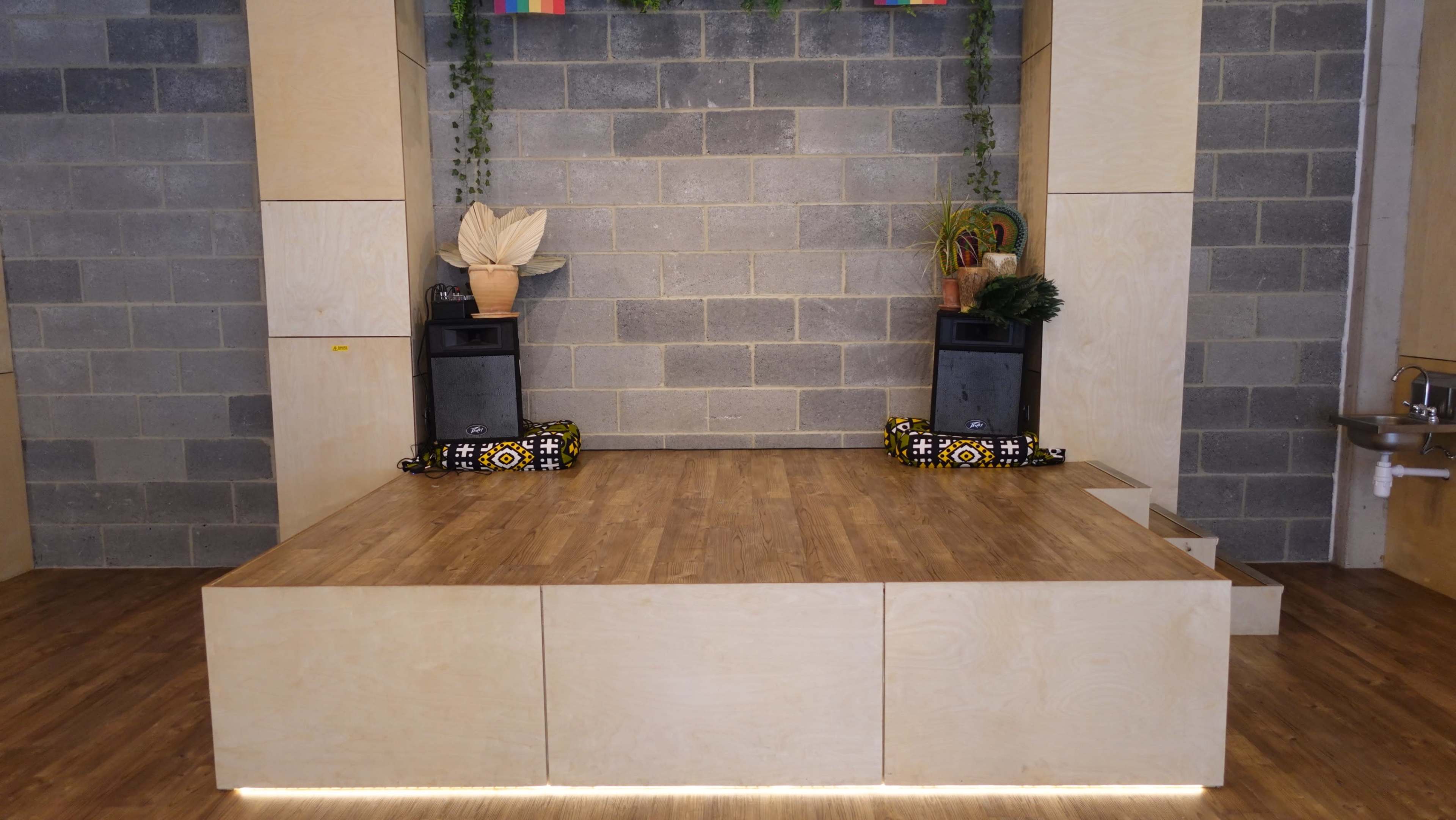 The image shows a wooden stage with two speakers on either side, set against a gray block wall with greenery and decorative items above.
