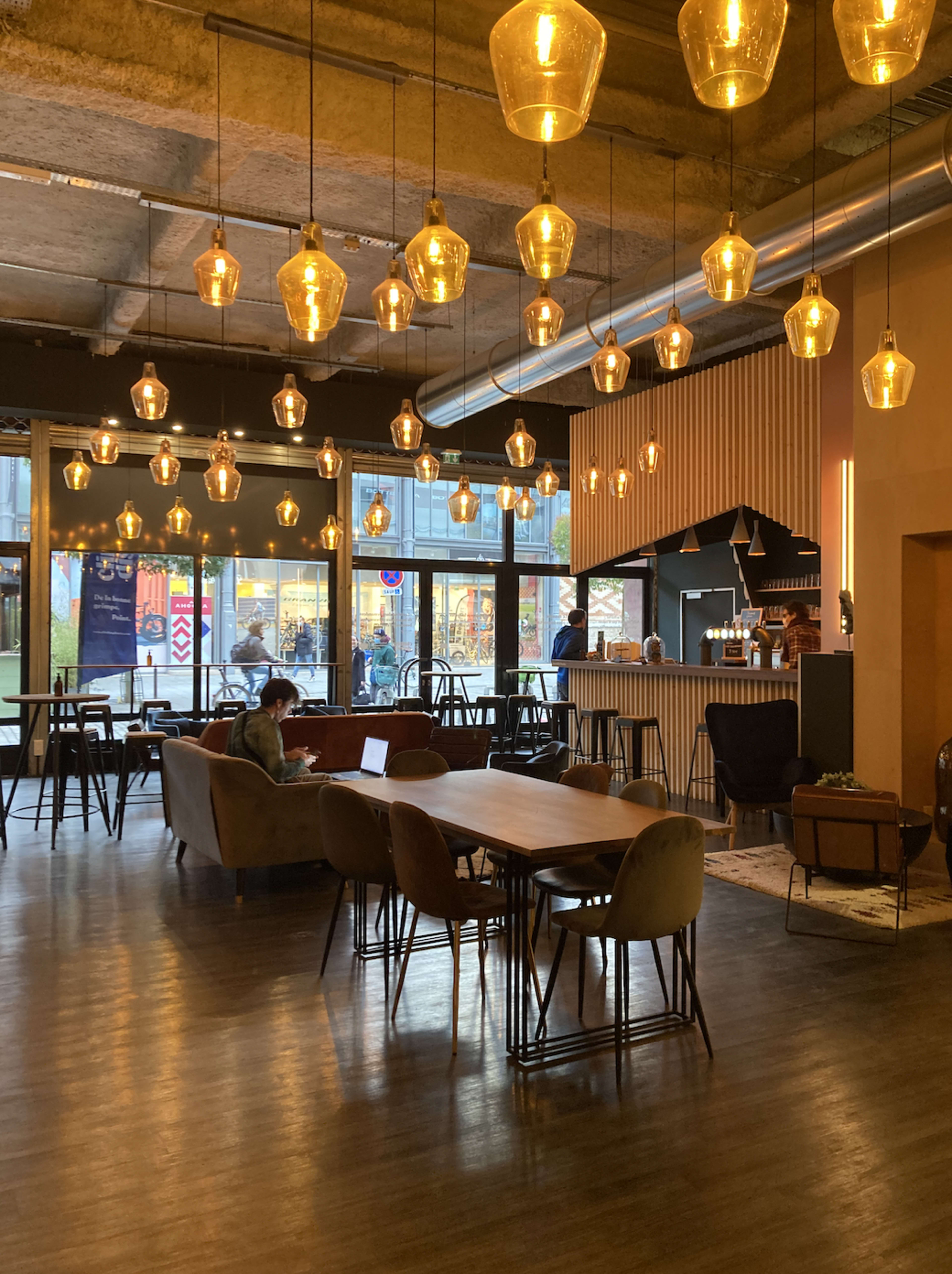 A well-lit cafe features hanging pendant lights, a mix of tables and seating, and large windows showcasing an outdoor view.