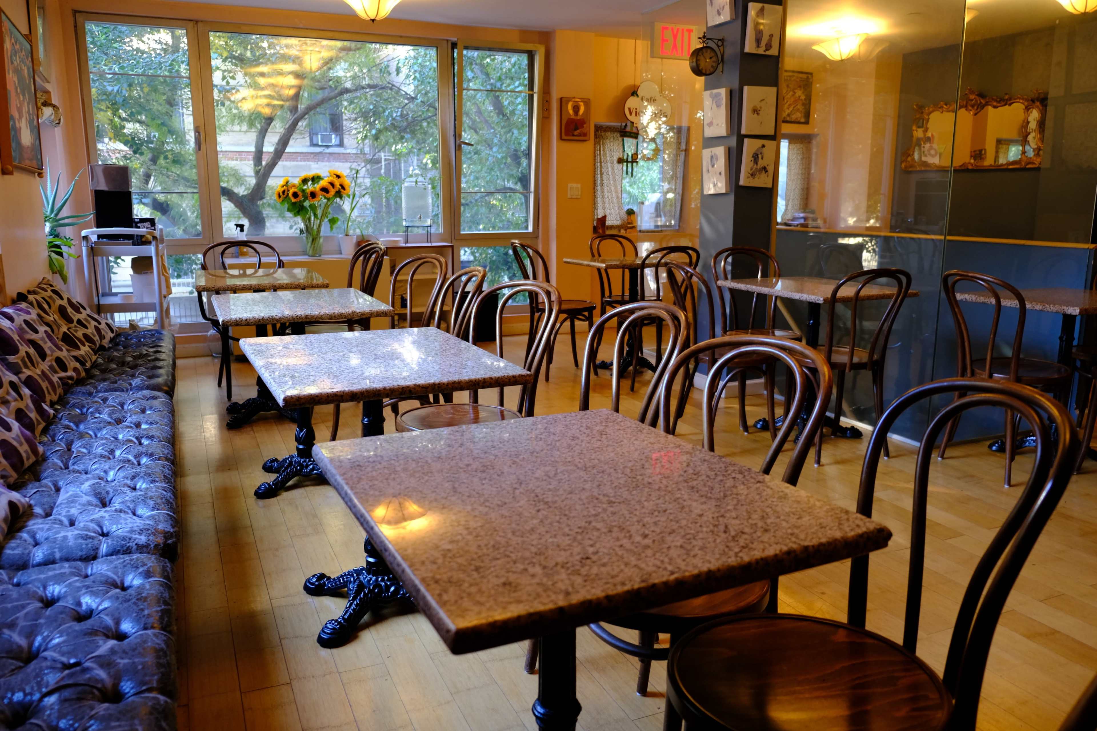 A cozy café with several marble-topped tables and wooden chairs arranged around a seating area with a cushioned sofa, illuminated by natural light through large windows.
