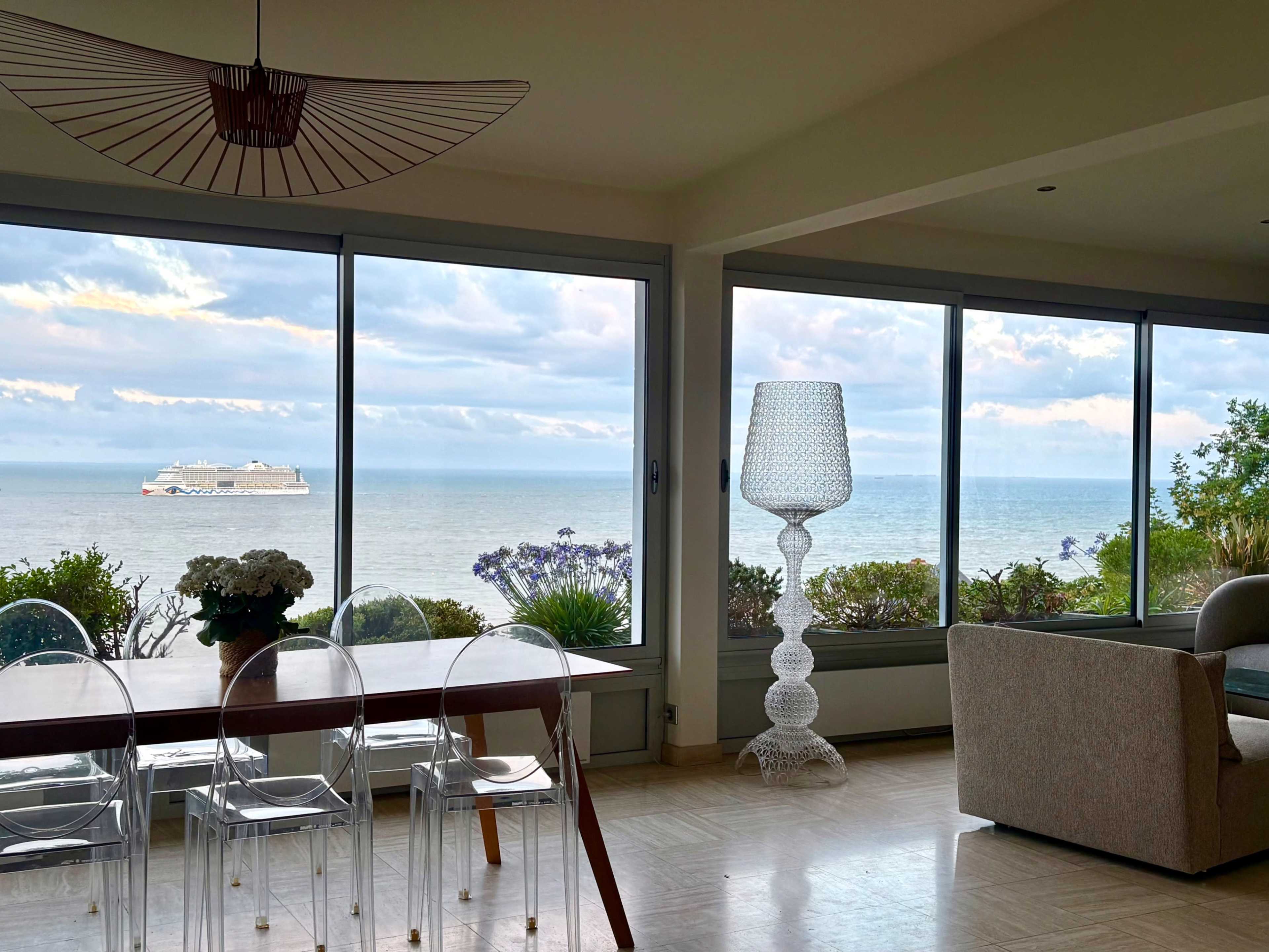 The image shows a modern interior with large windows overlooking a seaside view, featuring a ship in the distance and a large decorative lamp next to a dining table.