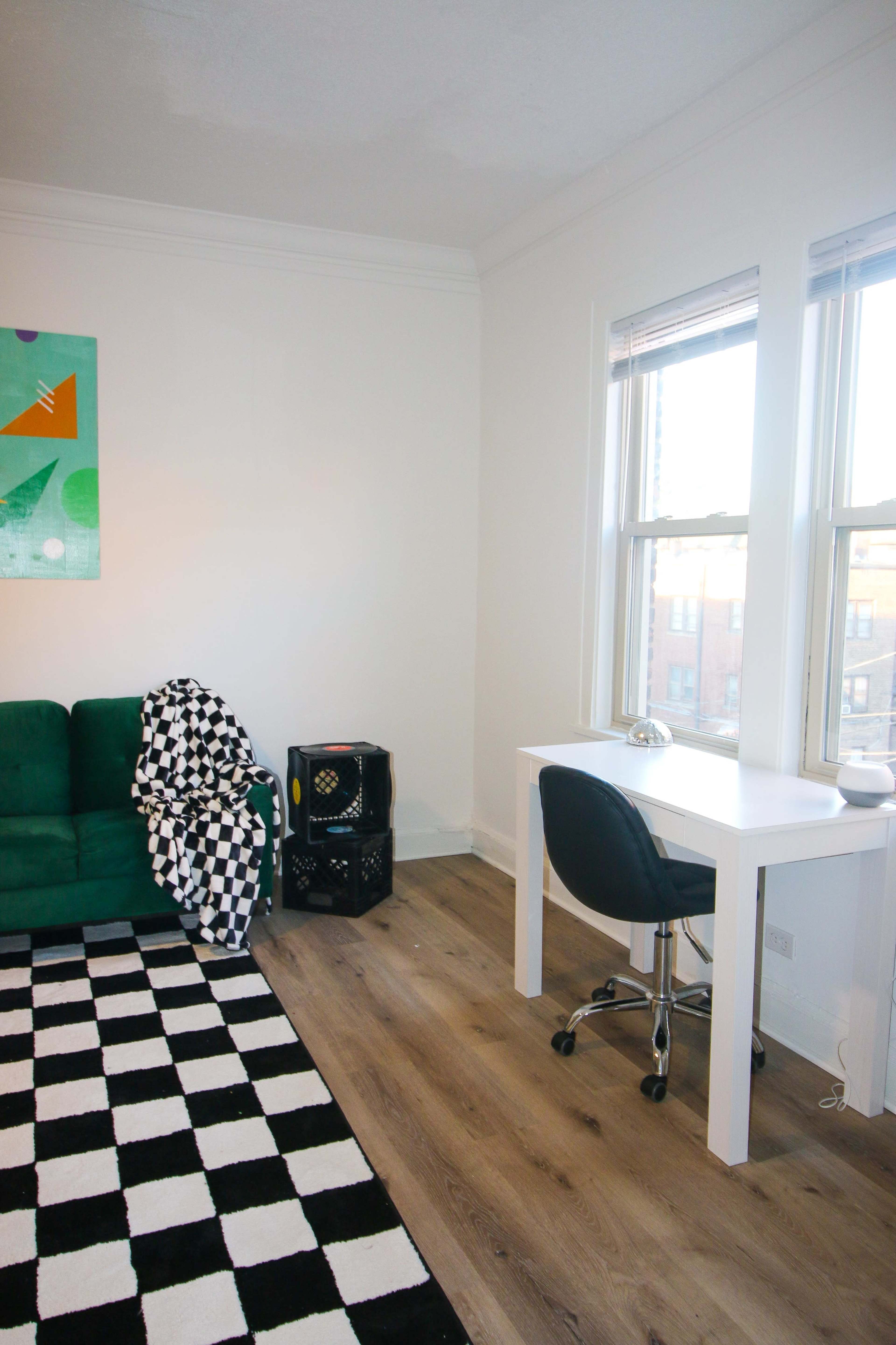 A room features a green couch with a black and white checkered blanket, a black and white patterned rug, and a white desk with a chair next to large windows.