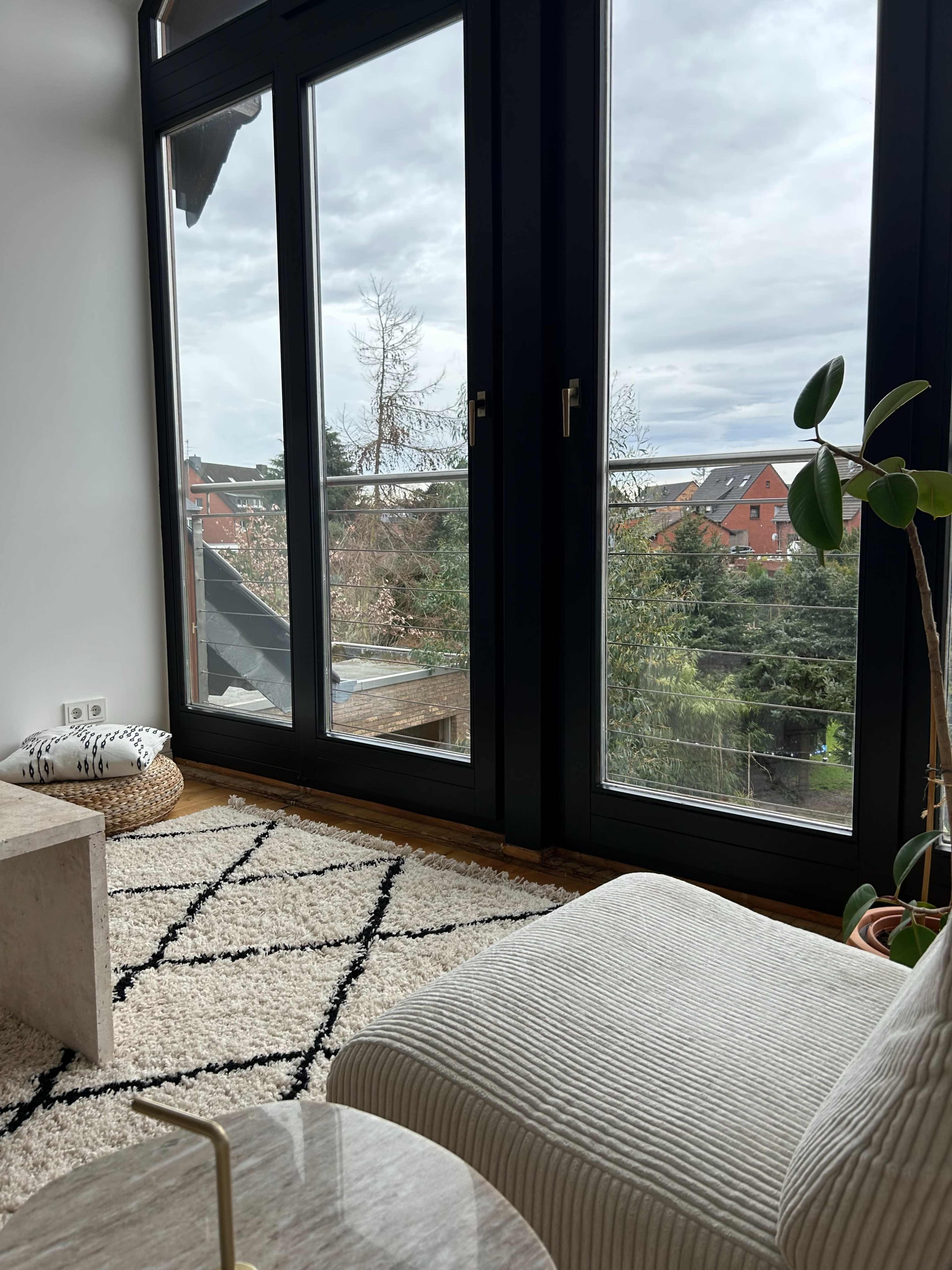 A cozy living room features a plush sofa, a coffee table, and large windows overlooking a green landscape and nearby buildings.
