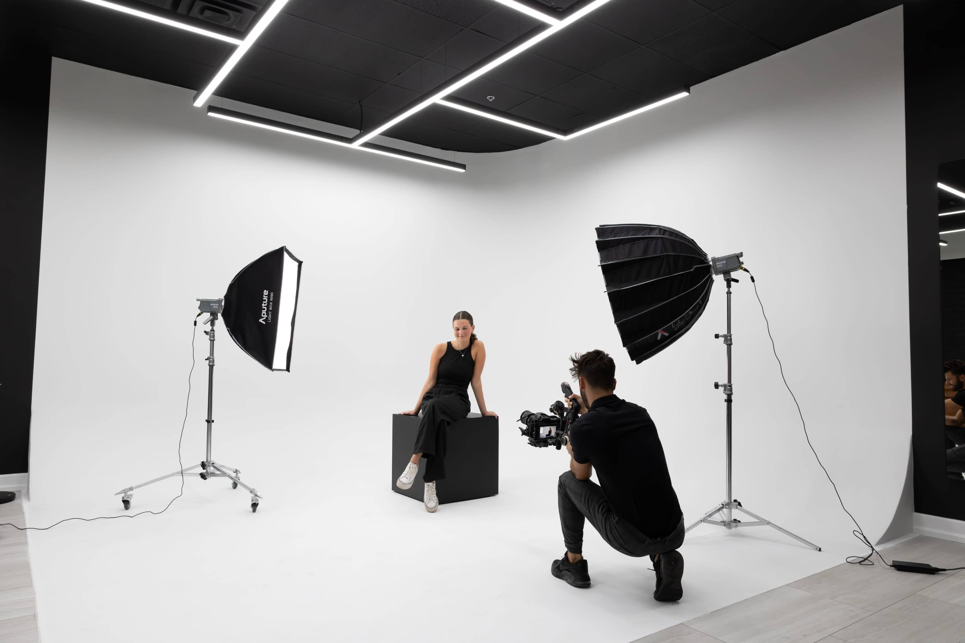 A photographer captures a model sitting on a black cube in a studio with a white backdrop and two softbox lights.