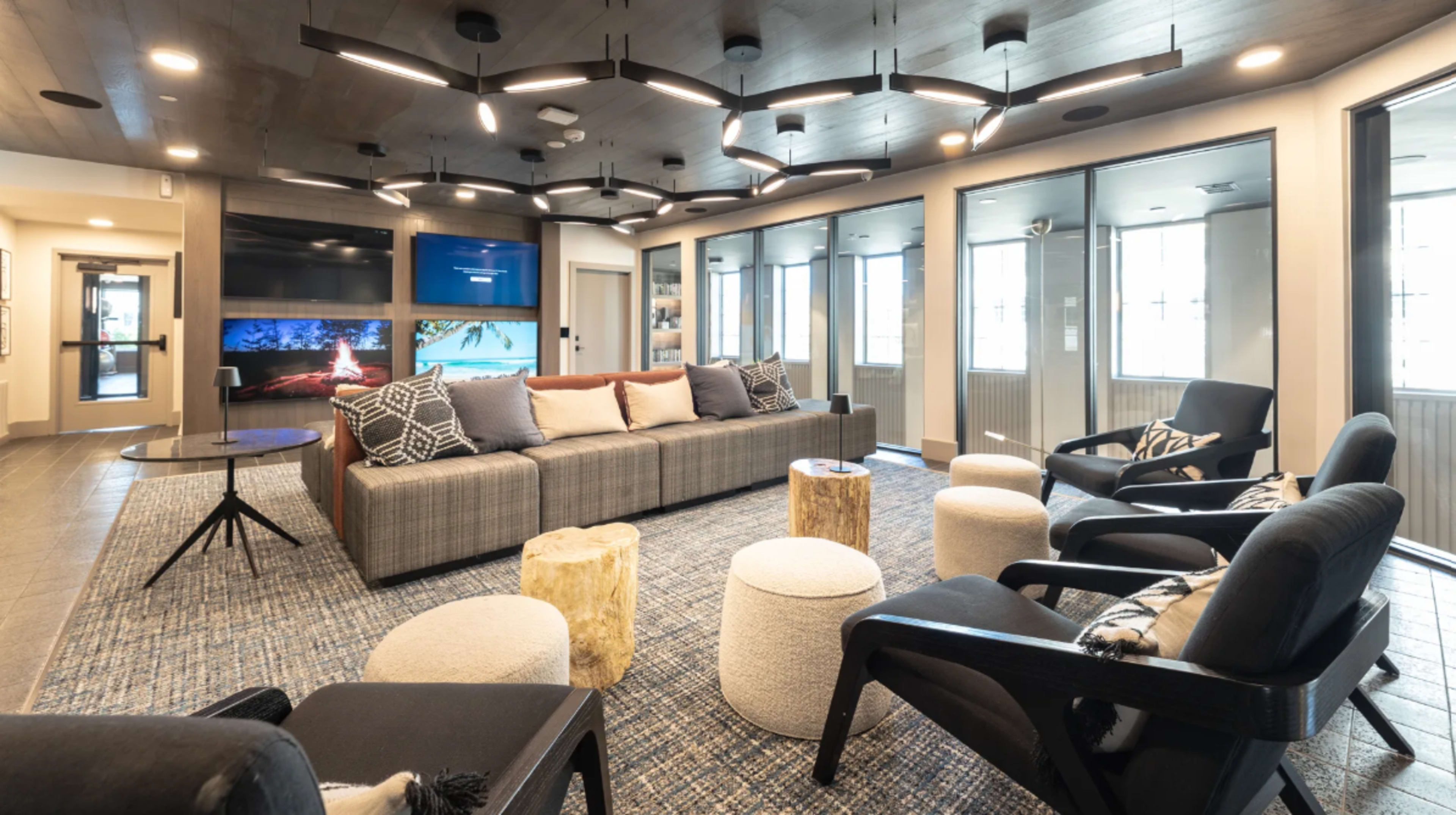 The image shows a modern lounge area with a sectional sofa, several chairs, a round table, and a large television mounted on the wall, illuminated by unique light fixtures.