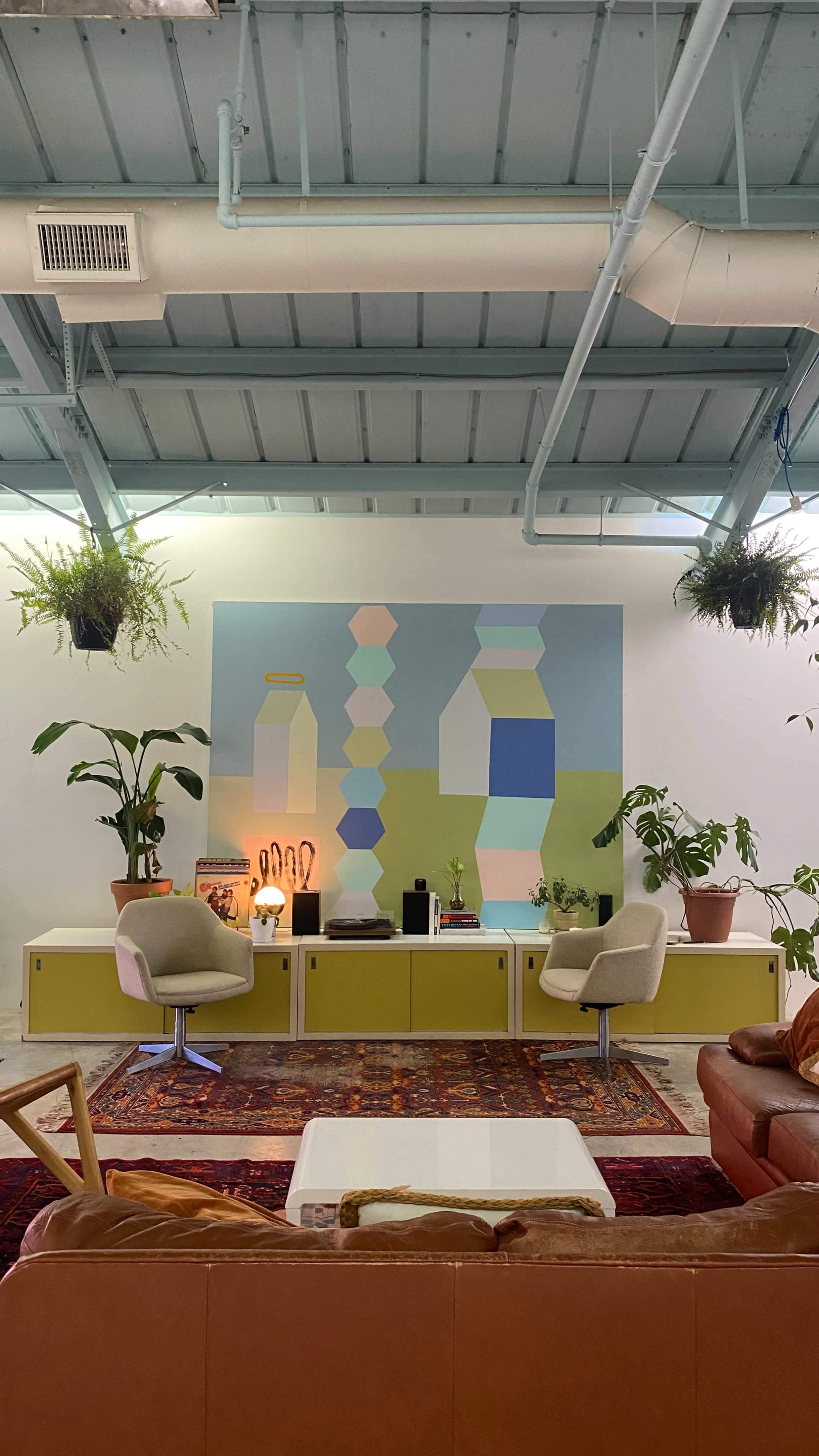 A modern living room features a mural of abstract shapes on the wall, with plants and contemporary furniture arranged around a coffee table.