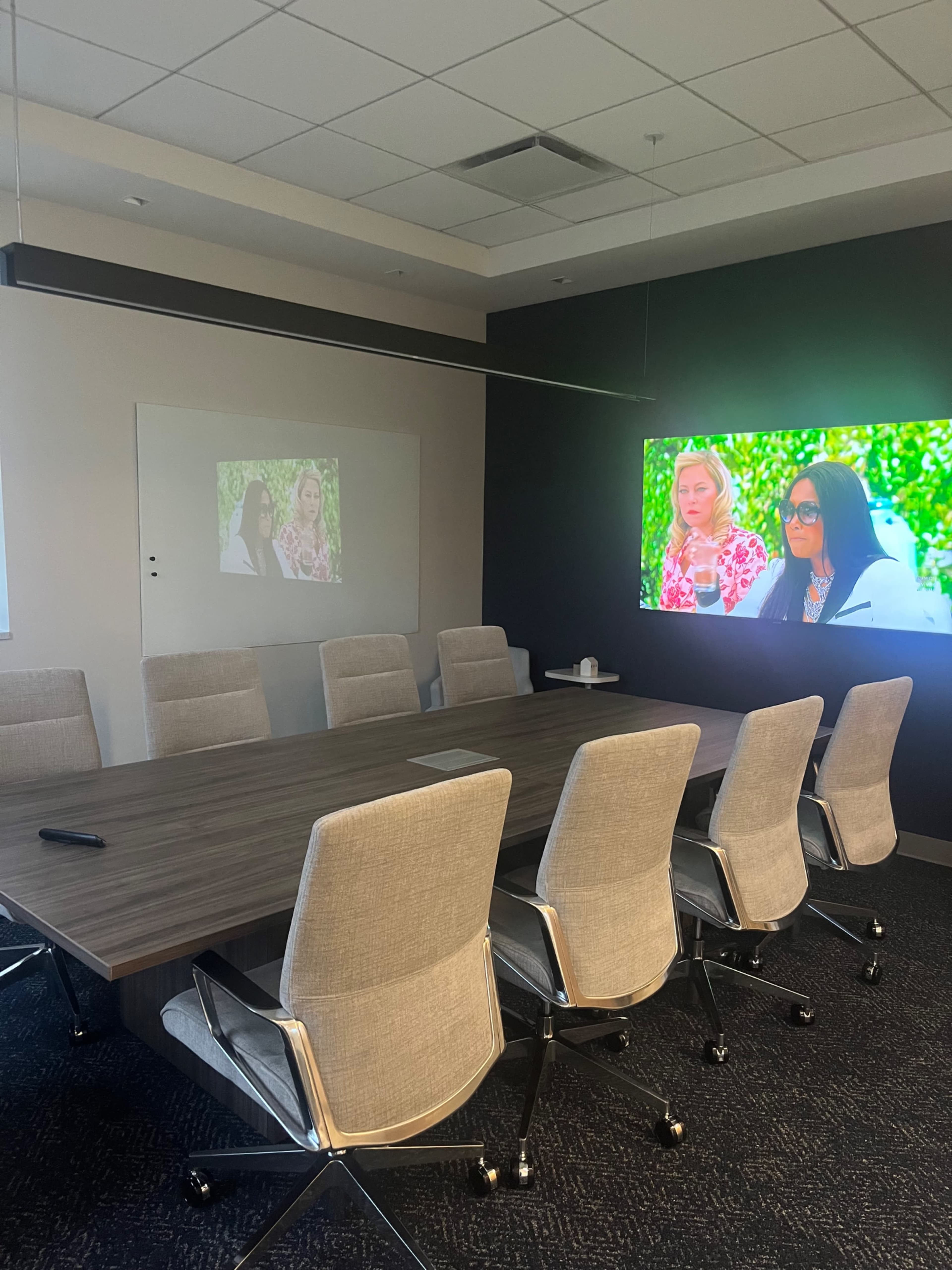 A modern conference room features a long wooden table, ergonomic chairs, and two screens displaying images on a dark wall.