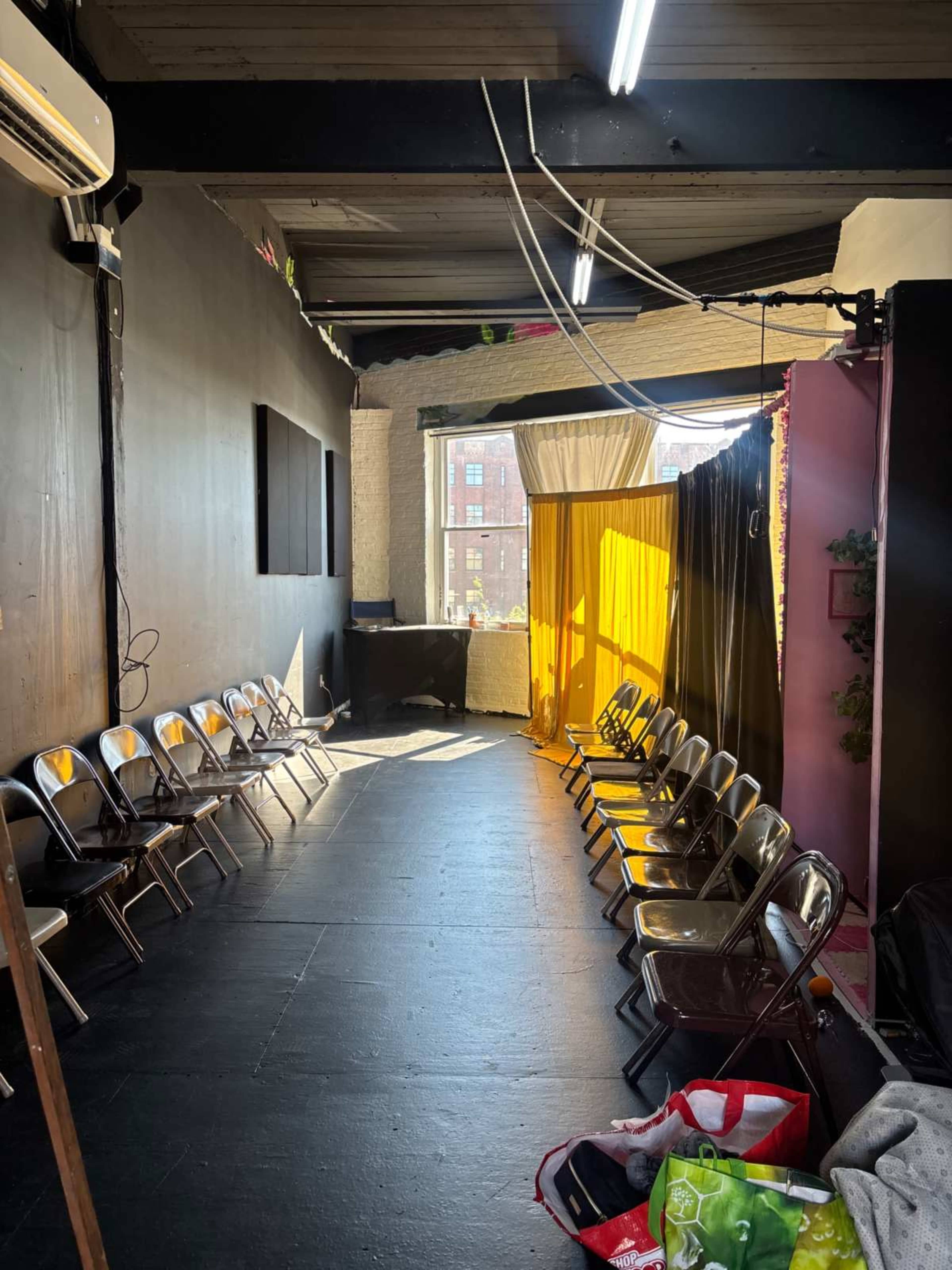 The image shows a narrow, well-lit room with folding chairs lined up along both sides and a section with draped curtains at one end.