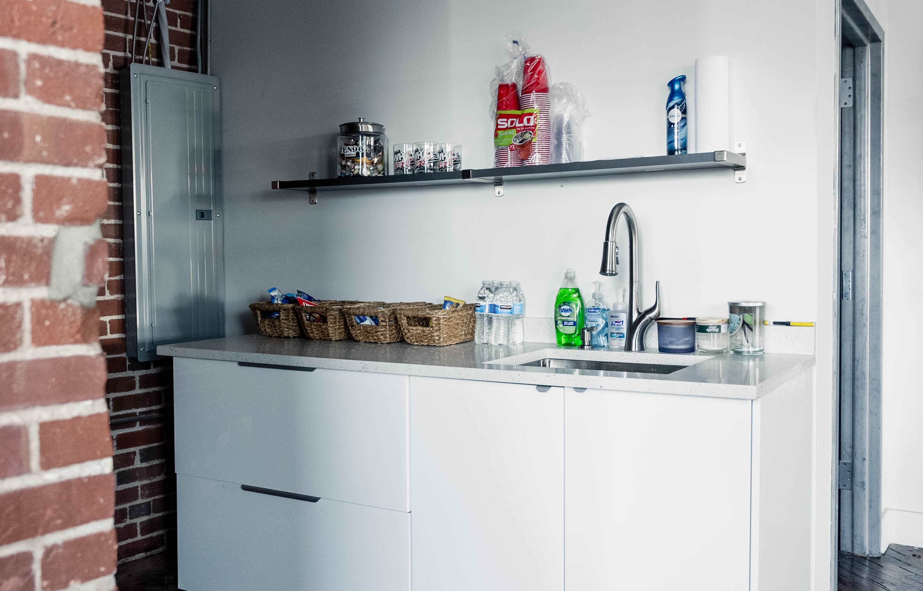 The image shows a modern kitchen area with a concrete countertop, a sink, and shelves above displaying various household items and cleaning supplies.