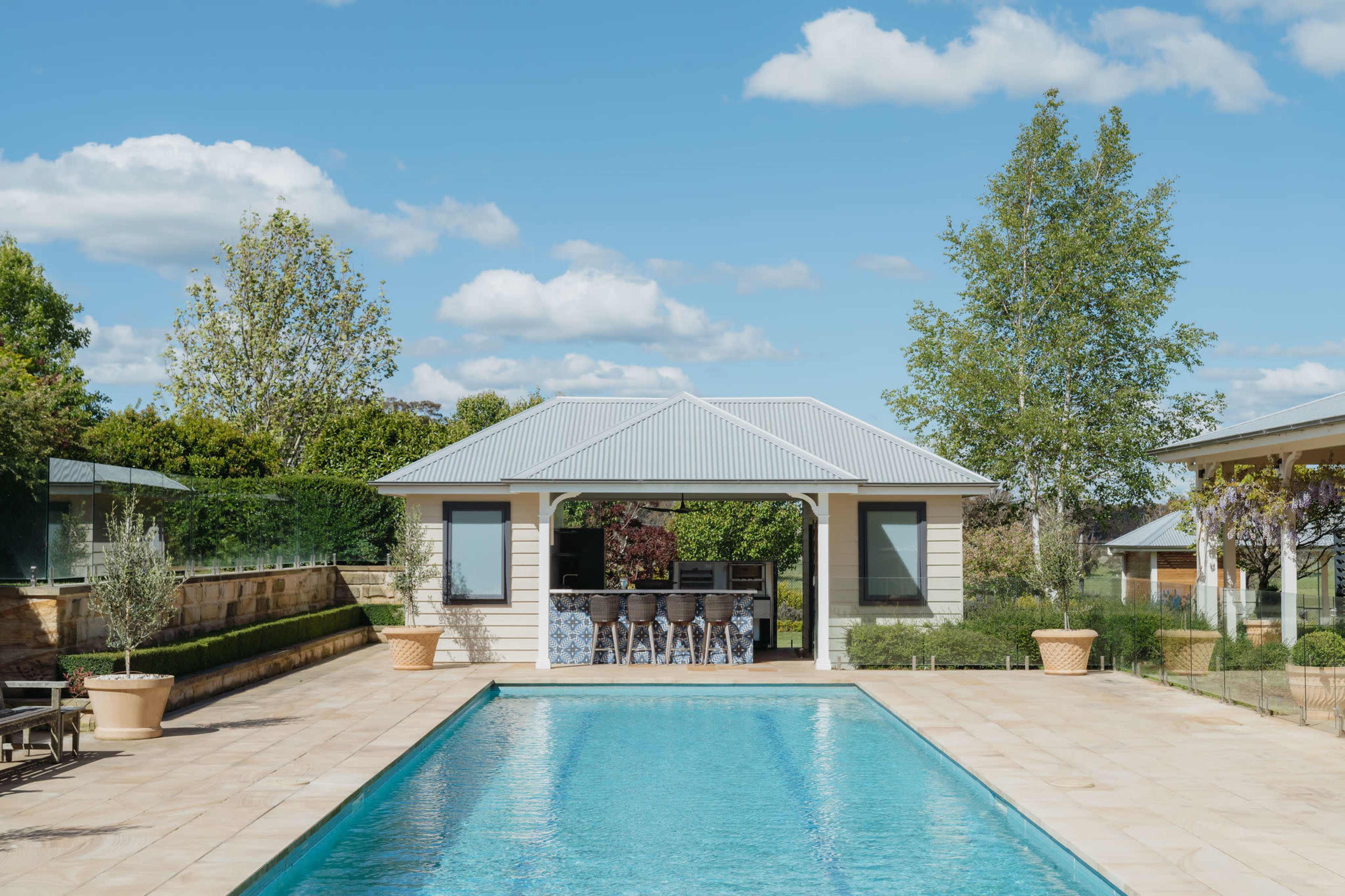 A rectangular swimming pool is set between a modern outdoor kitchen and a seated area, surrounded by landscaped gardens under a blue sky with scattered clouds.