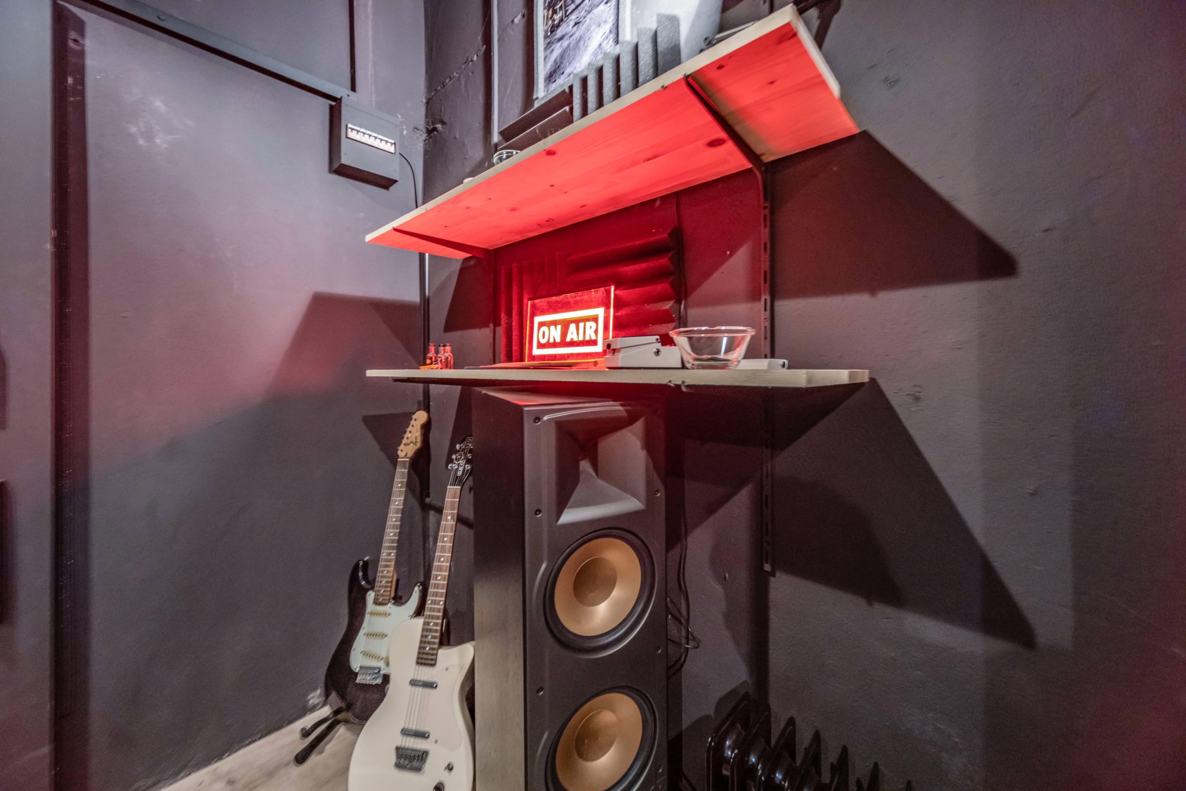 A corner of a room featuring two guitars, a pair of large speakers, a bowl, and a red "ON AIR" sign on a shelf against a dark wall.