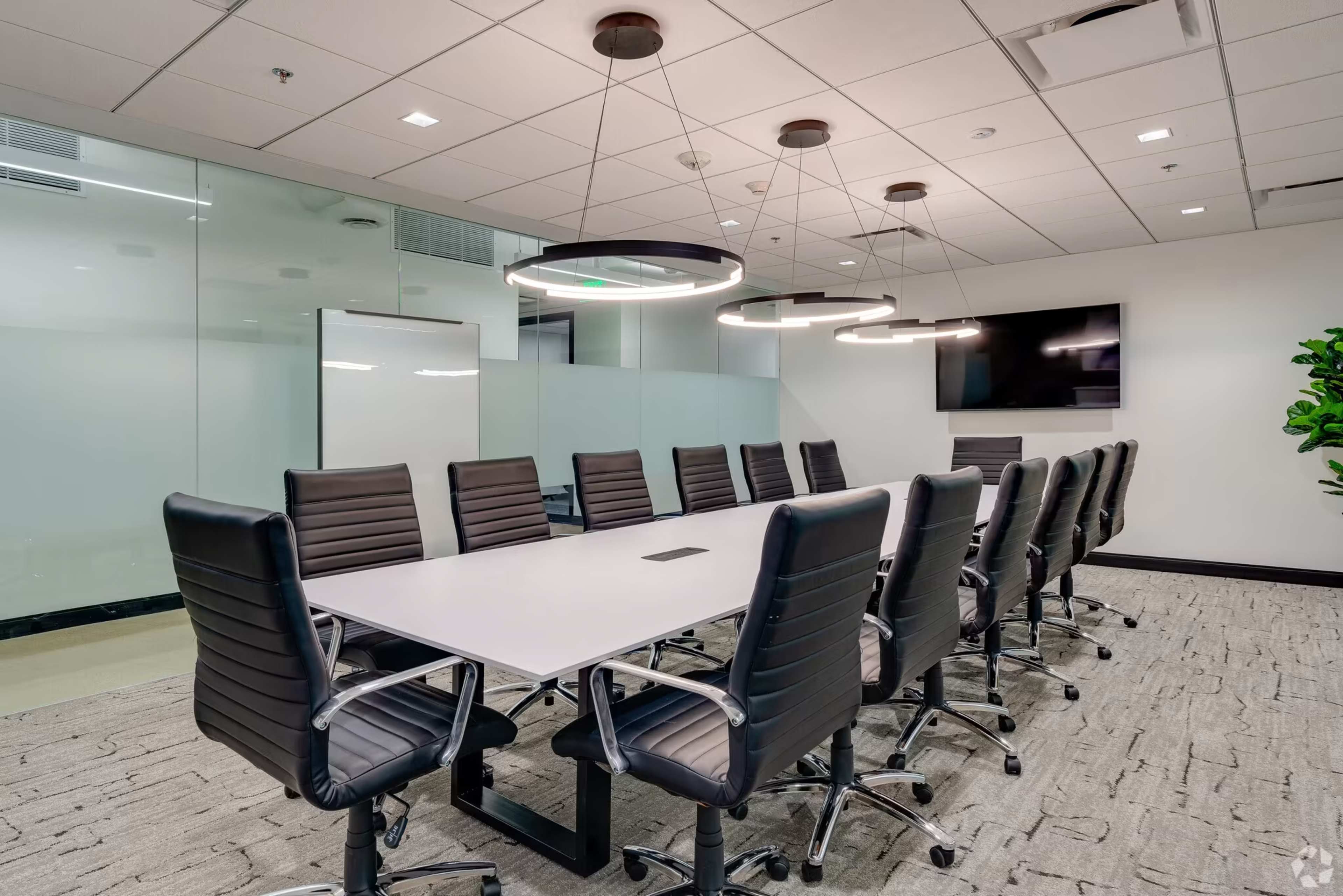 A modern conference room features a large table surrounded by black ergonomic chairs, with glass walls and circular light fixtures.