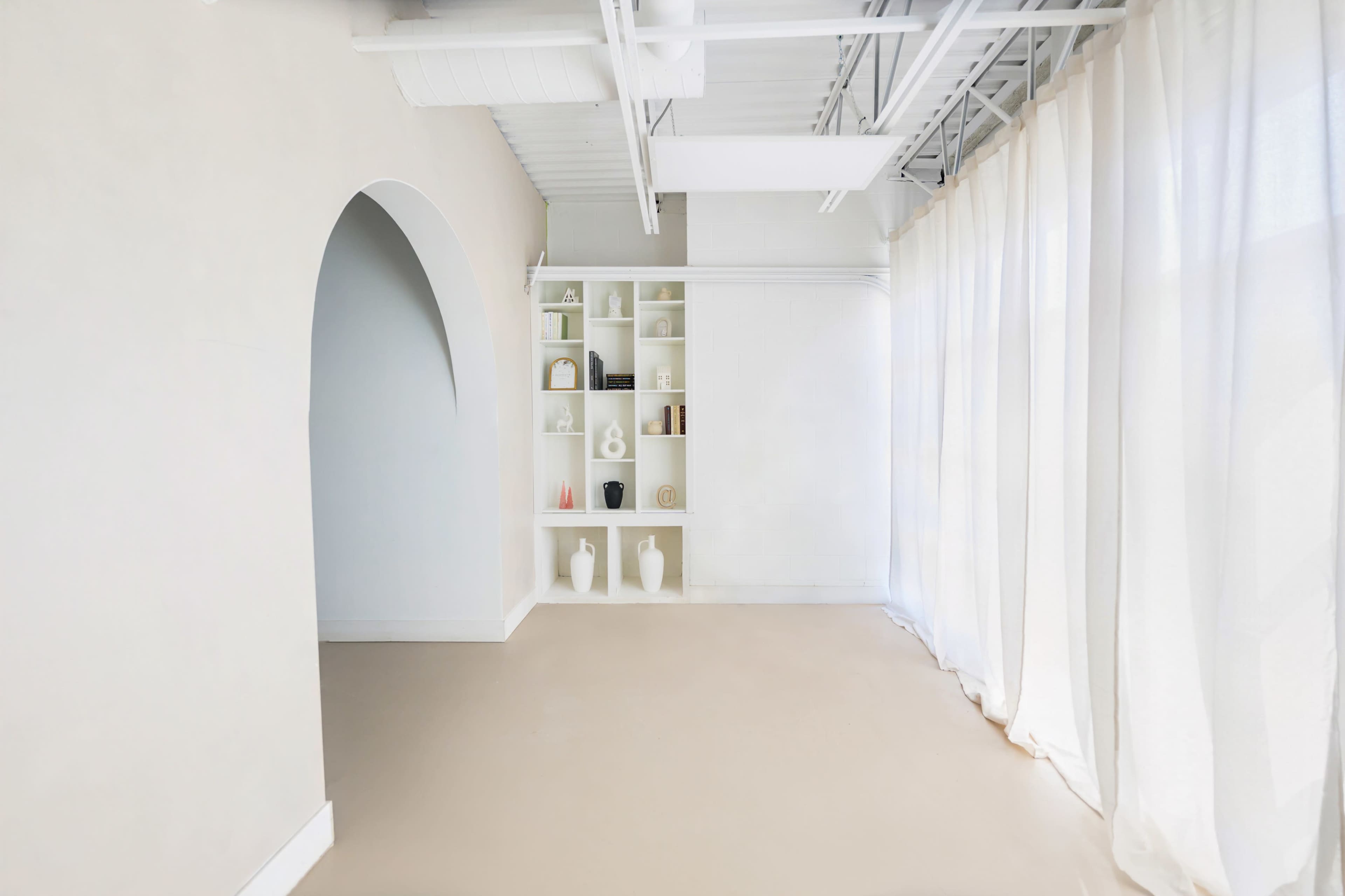 The image shows a minimalistic interior space with light-colored walls, a built-in shelf displaying various decorative items, and sheer curtains covering a large window.