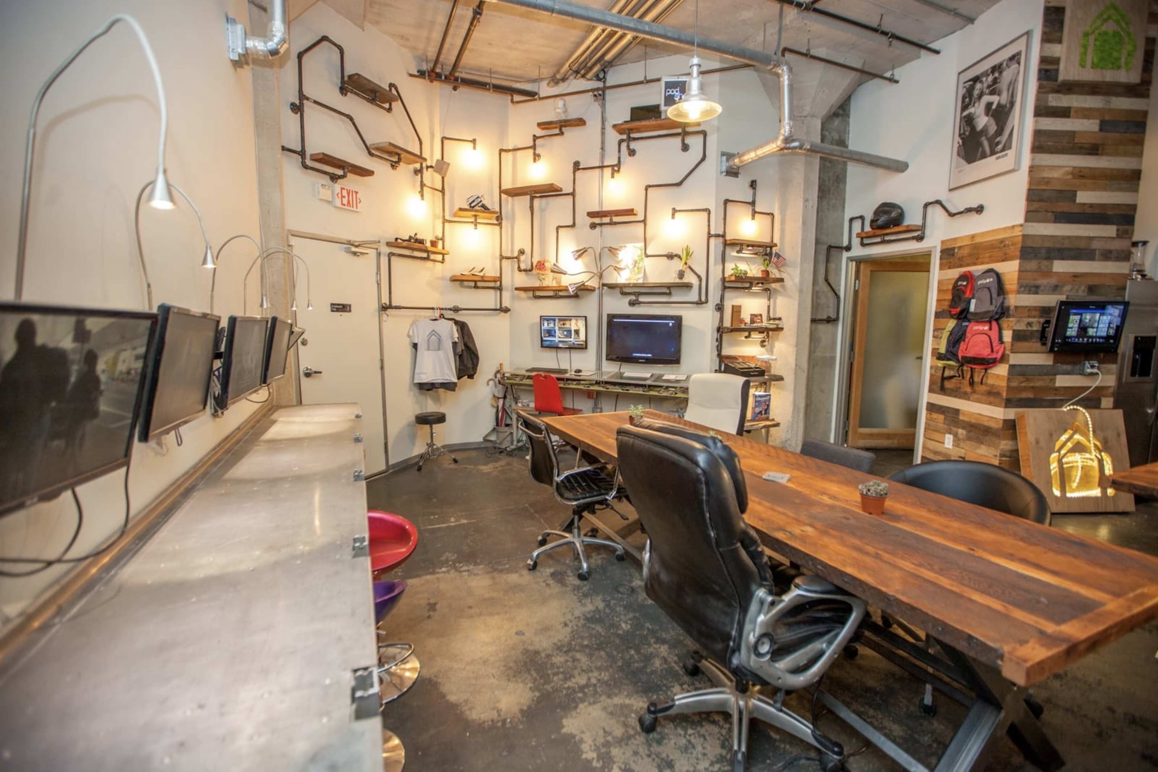 A modern office space featuring a wooden table, black leather chairs, multiple wall-mounted shelves with various items, and several computers on a stainless steel counter.