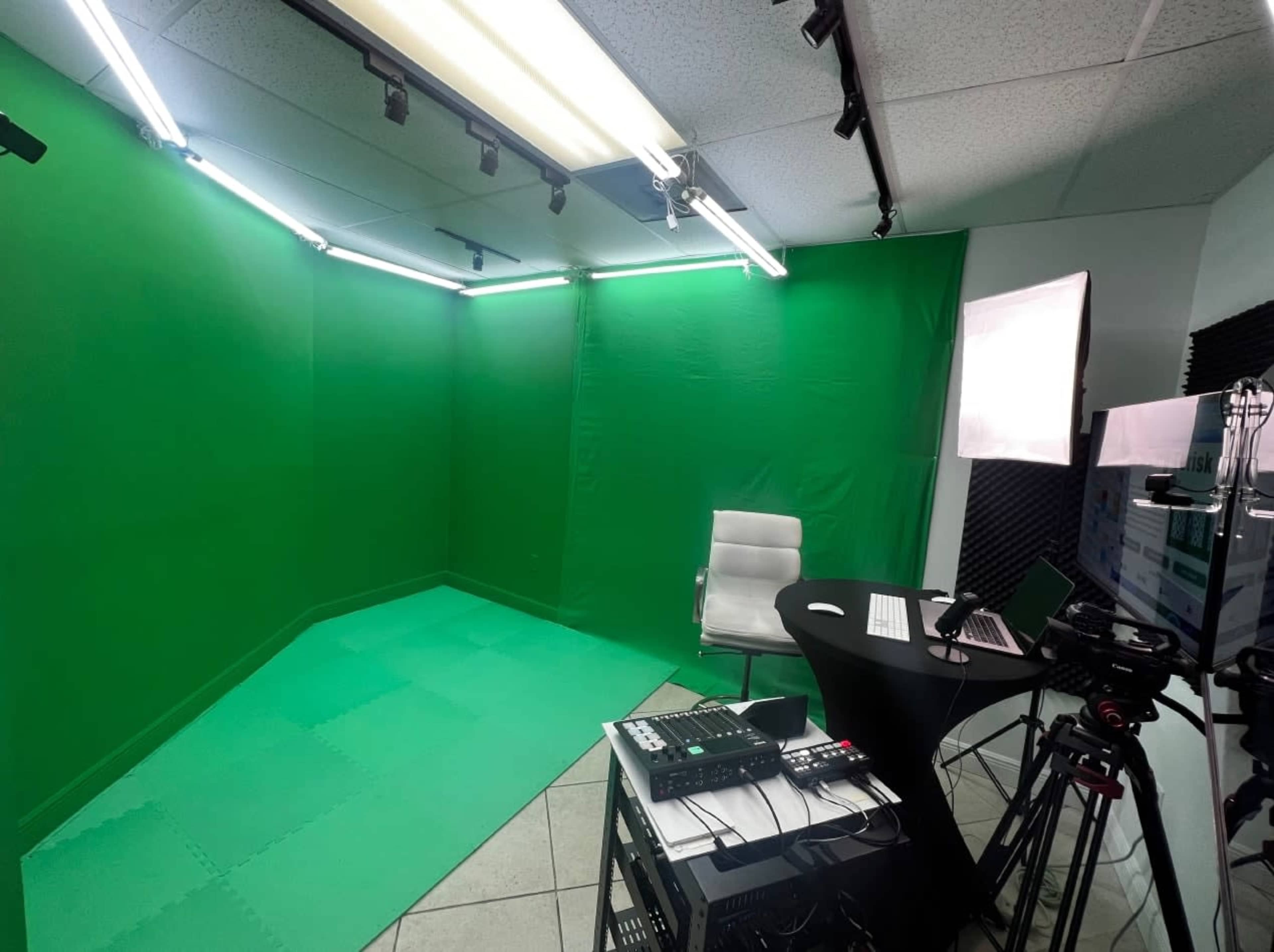 The image shows a green screen studio set up with a white chair, a black desk with a computer, and various recording equipment.