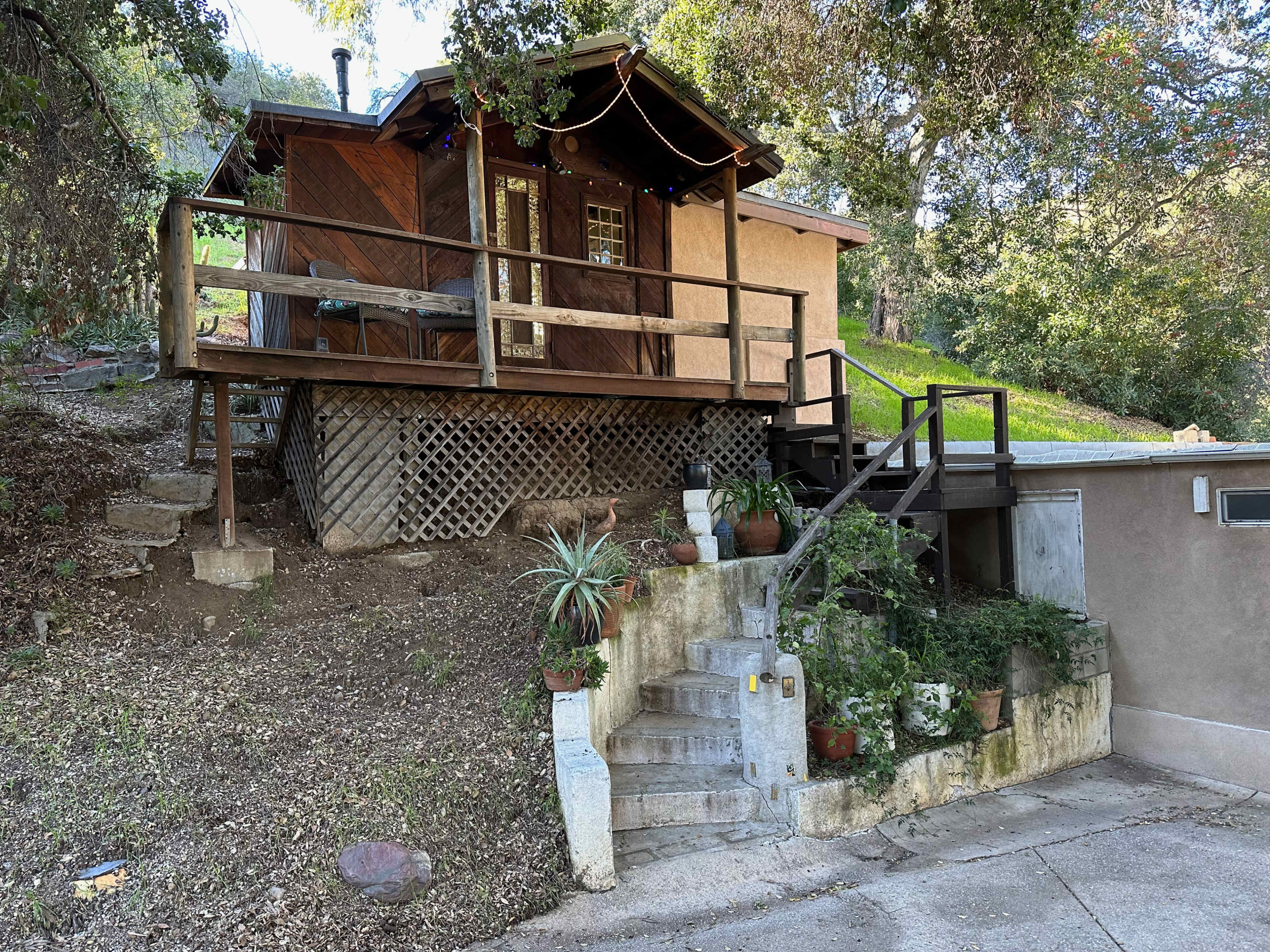 A small wooden cabin elevated on a hillside, with steps leading up to a porch and surrounded by greenery and potted plants.