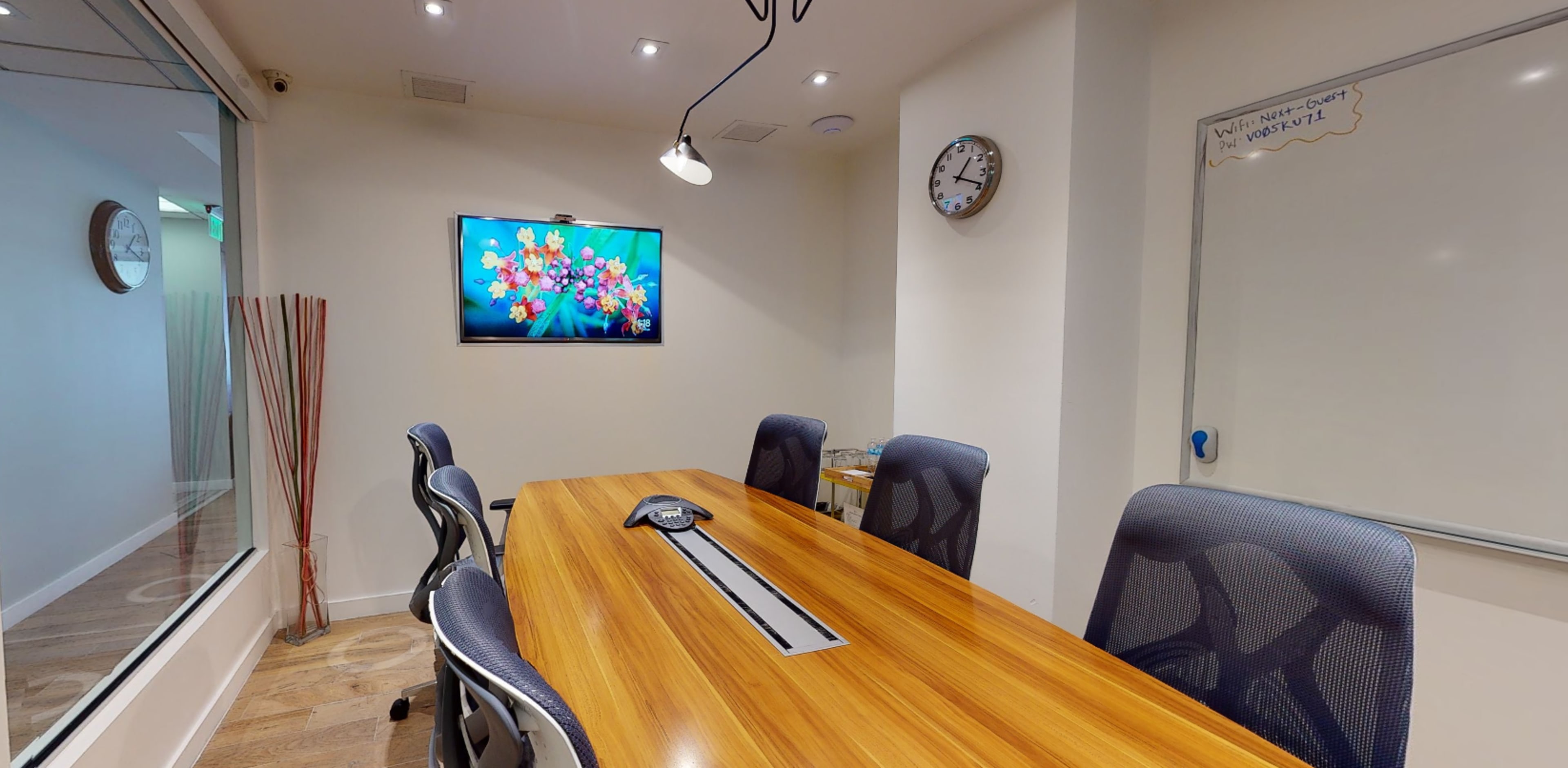 A conference room features a long wooden table with ergonomic chairs, a wall-mounted TV displaying colorful graphics, a clock, and a whiteboard.